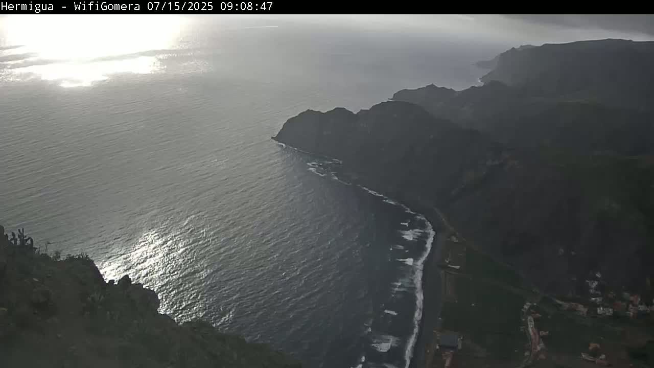 A high-angle view shows dark, rocky cliffs meeting a partly sunny ocean with small waves breaking along a shoreline.