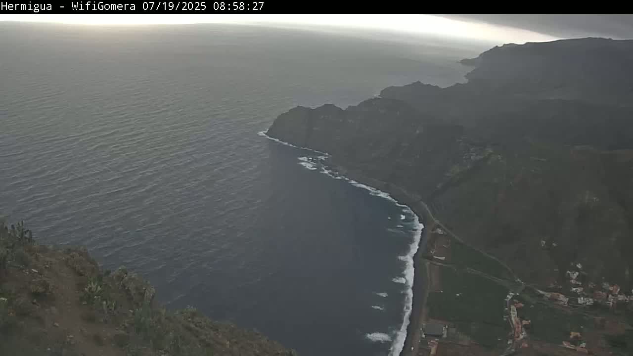 A high-angle view shows a dark, overcast sky over a rocky coastline where dark-colored land meets a dark-colored ocean with small waves.