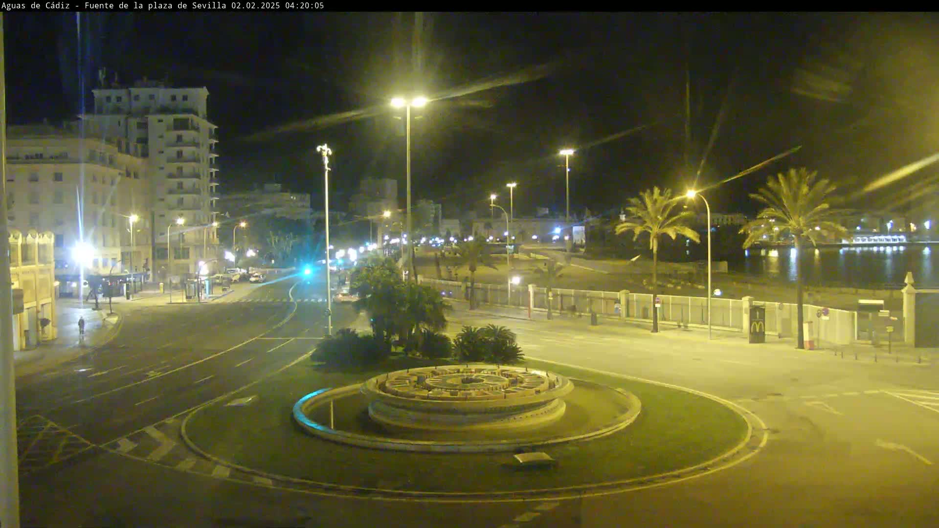 A nighttime view of a city roundabout with a circular fountain at its center, illuminated by streetlights, with palm trees and buildings visible in the background.
