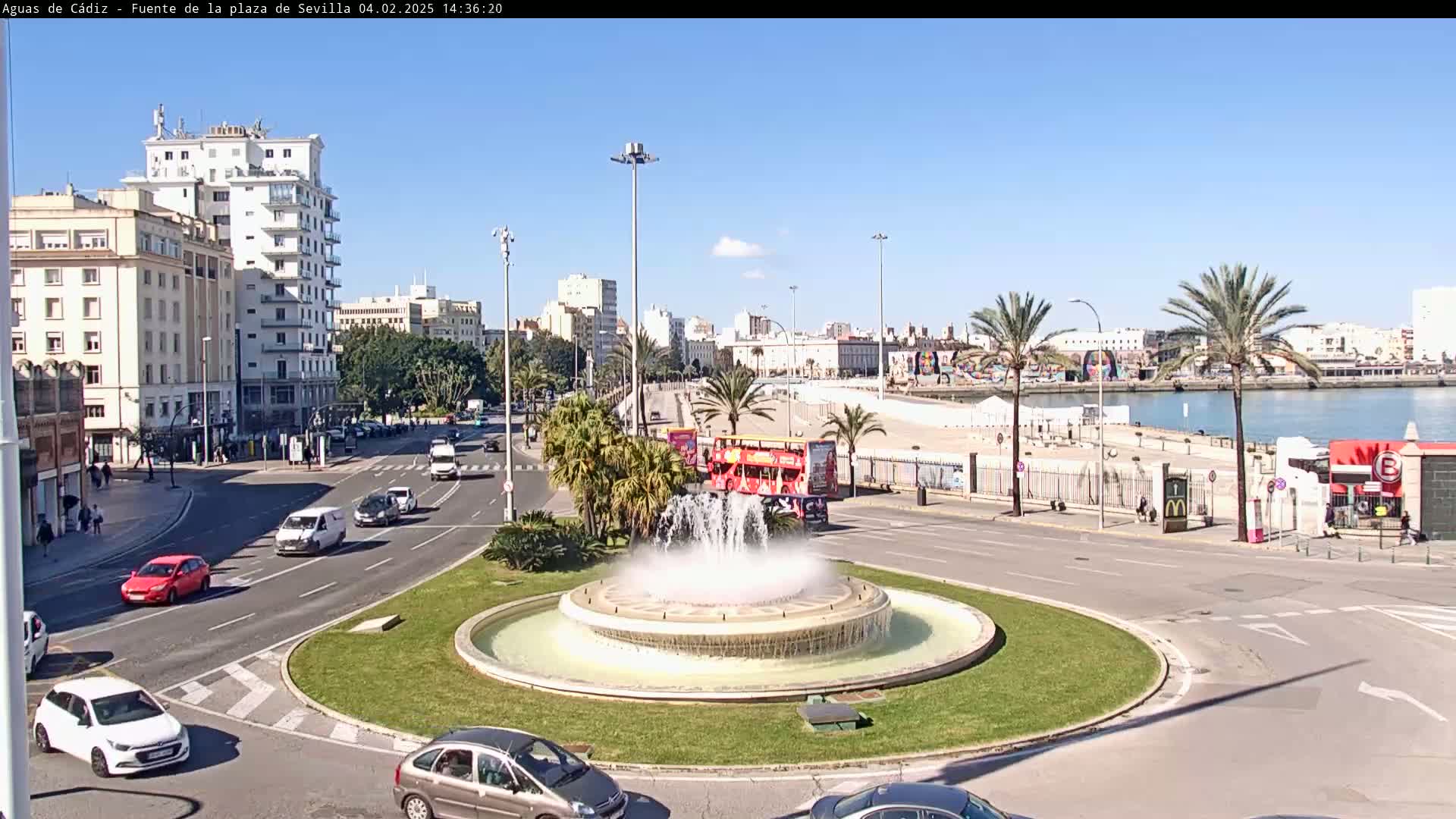 A traffic circle with a fountain in the center is surrounded by city buildings, palm trees, and a waterfront under sunny skies.