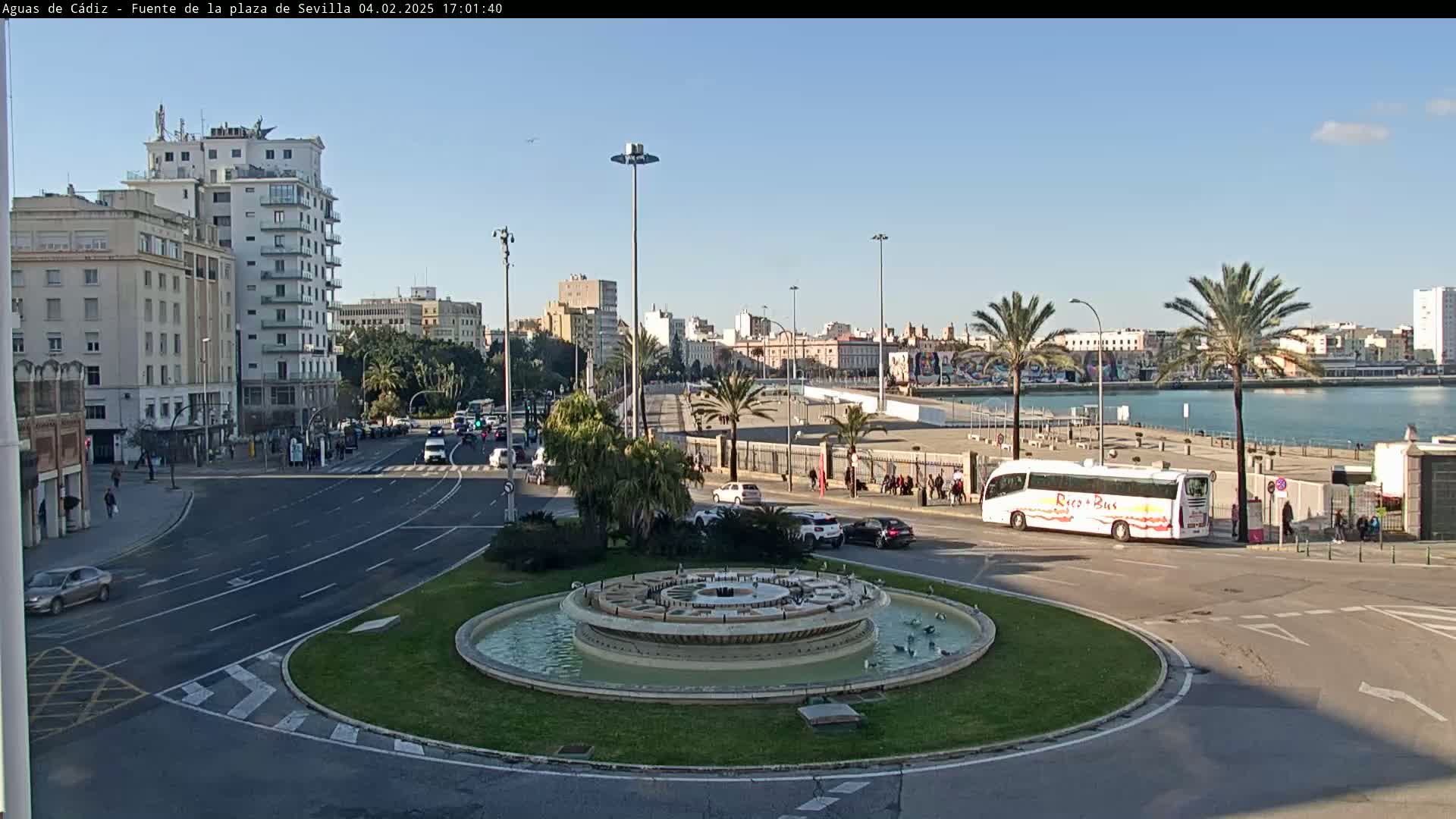 A city roundabout with a fountain in the center, surrounded by roads, buildings, and palm trees, under a clear, sunny sky.