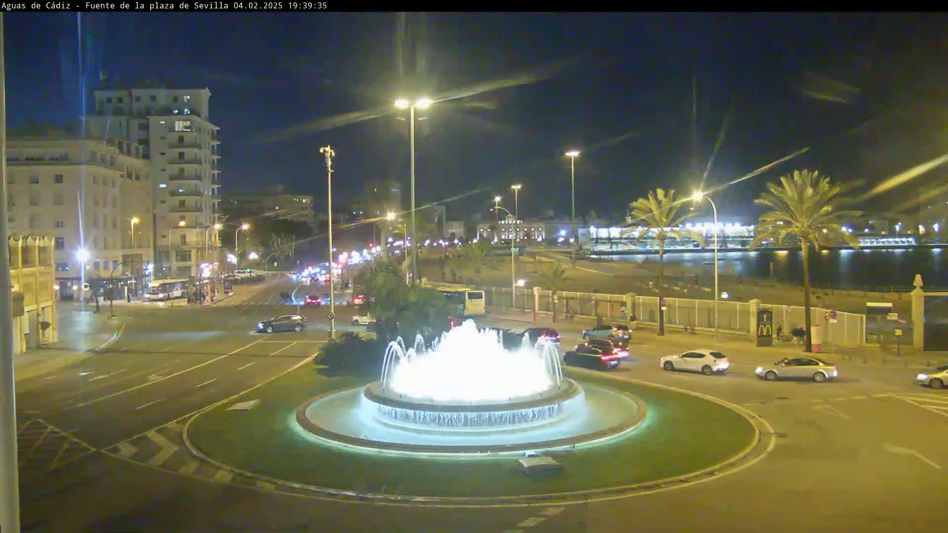 A nighttime view of a city roundabout featuring a lit fountain, with cars driving around it and buildings and palm trees visible in the background under a clear, dark sky.