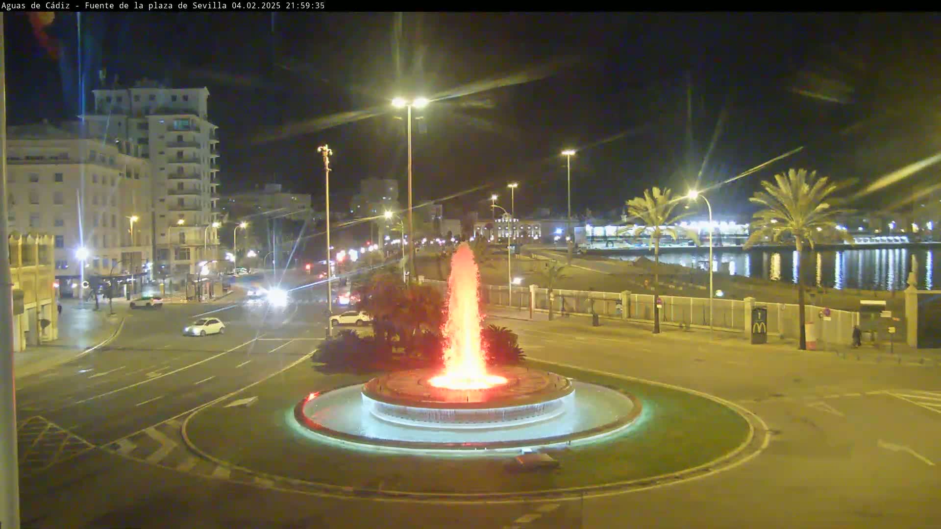 A nighttime view of a city square with a tall fountain spraying red water, cars on the road, and palm trees near a calm body of water in the background.