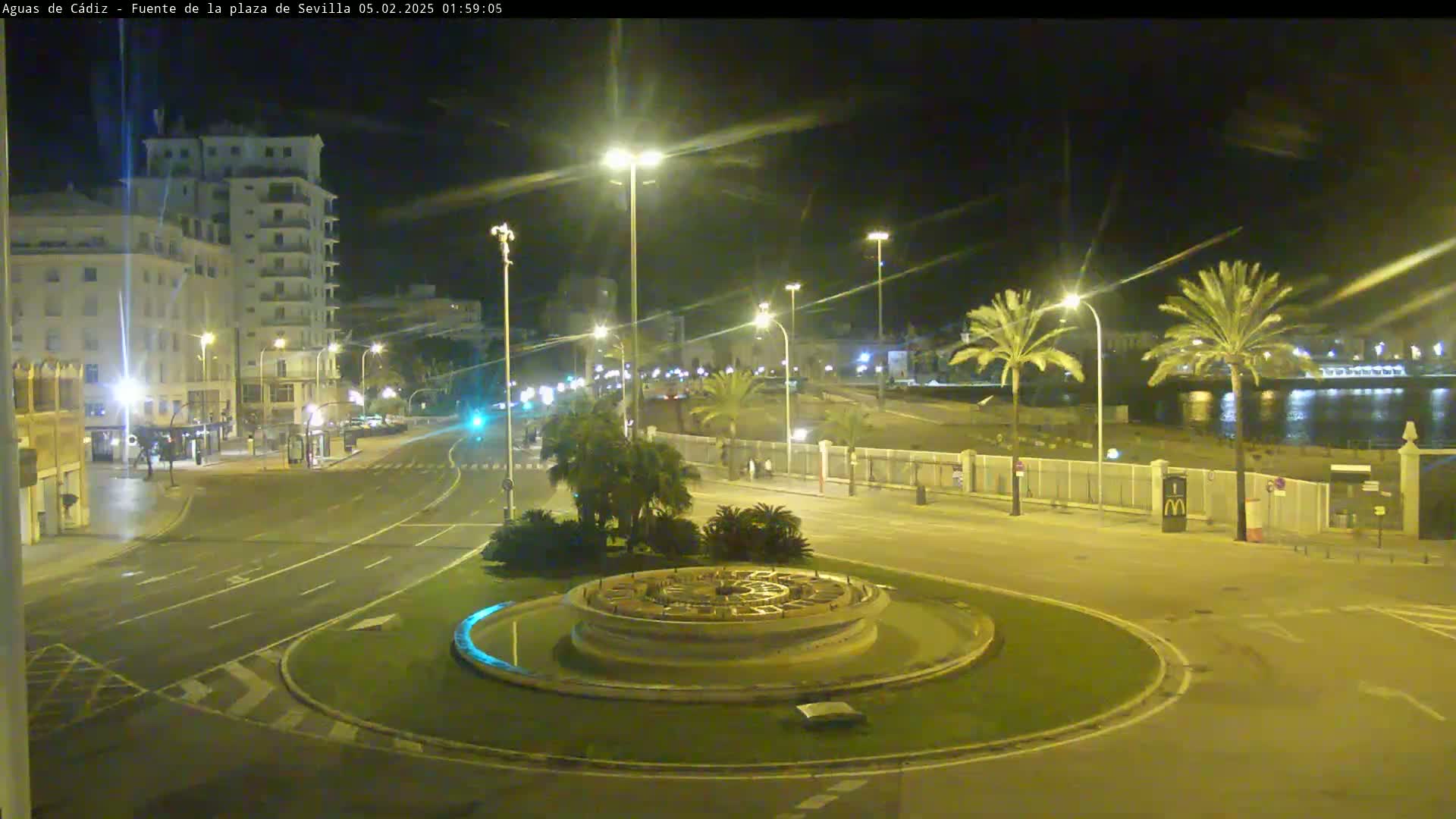 A nighttime view of a circular plaza with a fountain at its center, surrounded by palm trees, buildings, and roadways, near a body of water under clear skies.