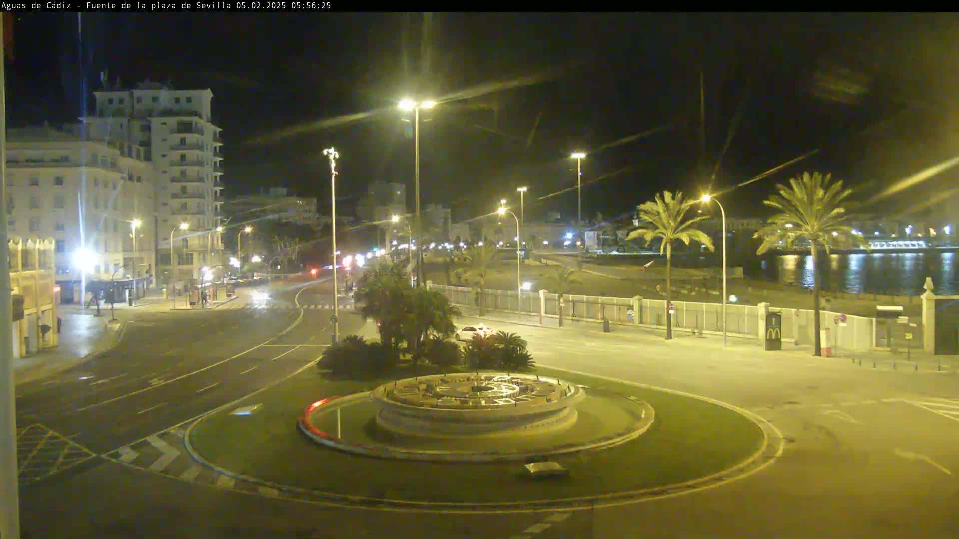 A nighttime view of a city roundabout with a fountain at its center,  illuminated by streetlights, near a waterfront with palm trees under a clear sky.