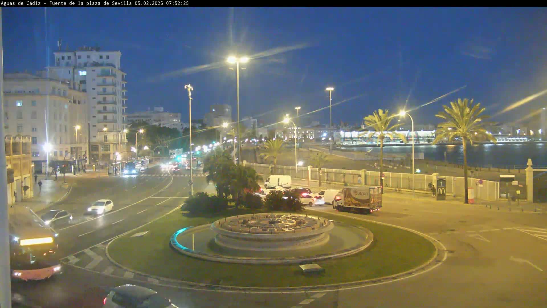 A nighttime view of a city roundabout with a fountain at its center, featuring traffic moving around it and a waterfront in the background under a twilight sky.