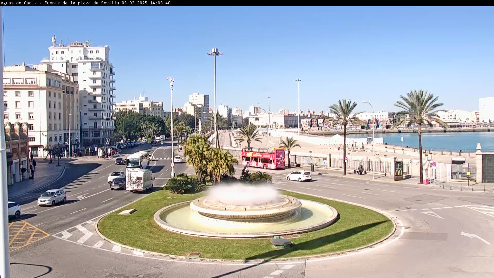 A circular fountain is in the foreground of a city street scene on a sunny day, with vehicles traveling on the road and palm trees lining a waterfront in the background.