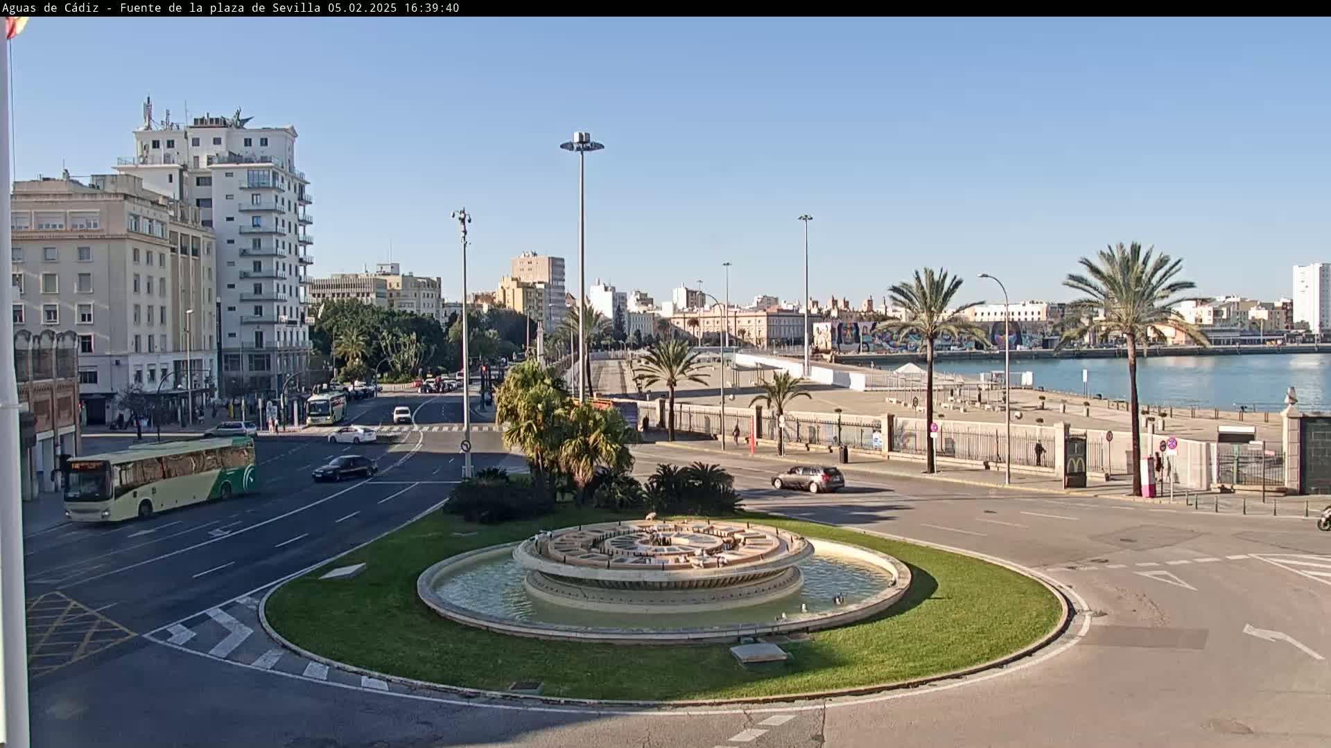 A circular fountain sits in the center of a traffic circle, with city buildings, palm trees, and a body of water visible in the background under a clear, sunny sky.