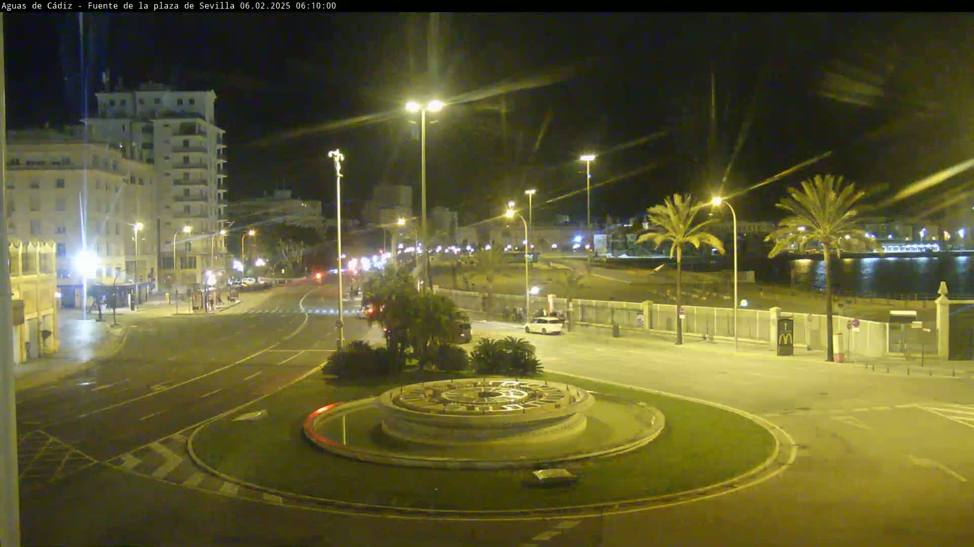 A nighttime view of a city roundabout with a fountain at its center, surrounded by palm trees, buildings, and roadways, under clear skies.