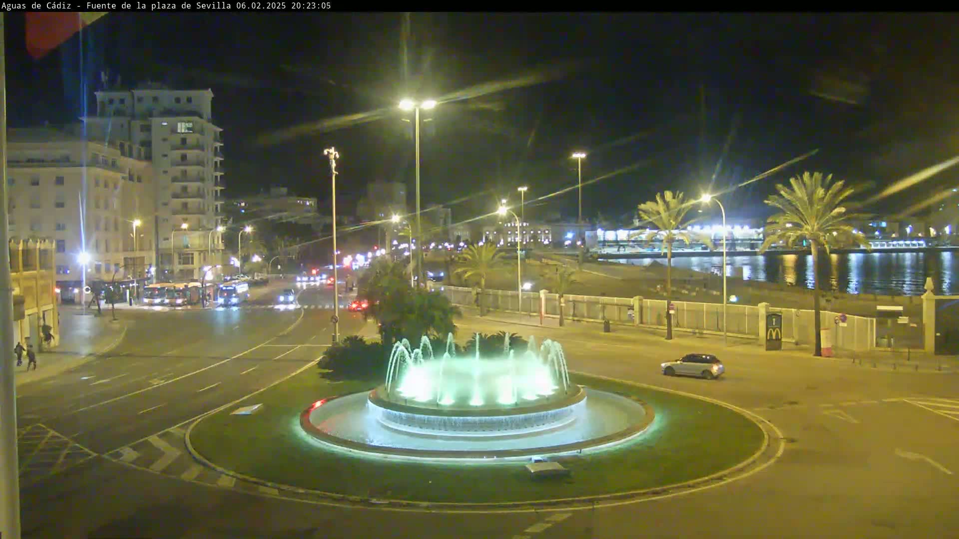 A nighttime view of a city square featuring a circular fountain with illuminated jets of water, surrounded by streets with cars and buildings, near a calm body of water under clear skies.