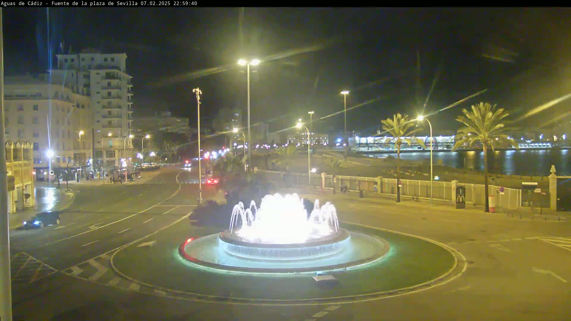 A nighttime view of a city square with a lit fountain at its center, surrounded by roads and buildings, with calm water visible in the background.