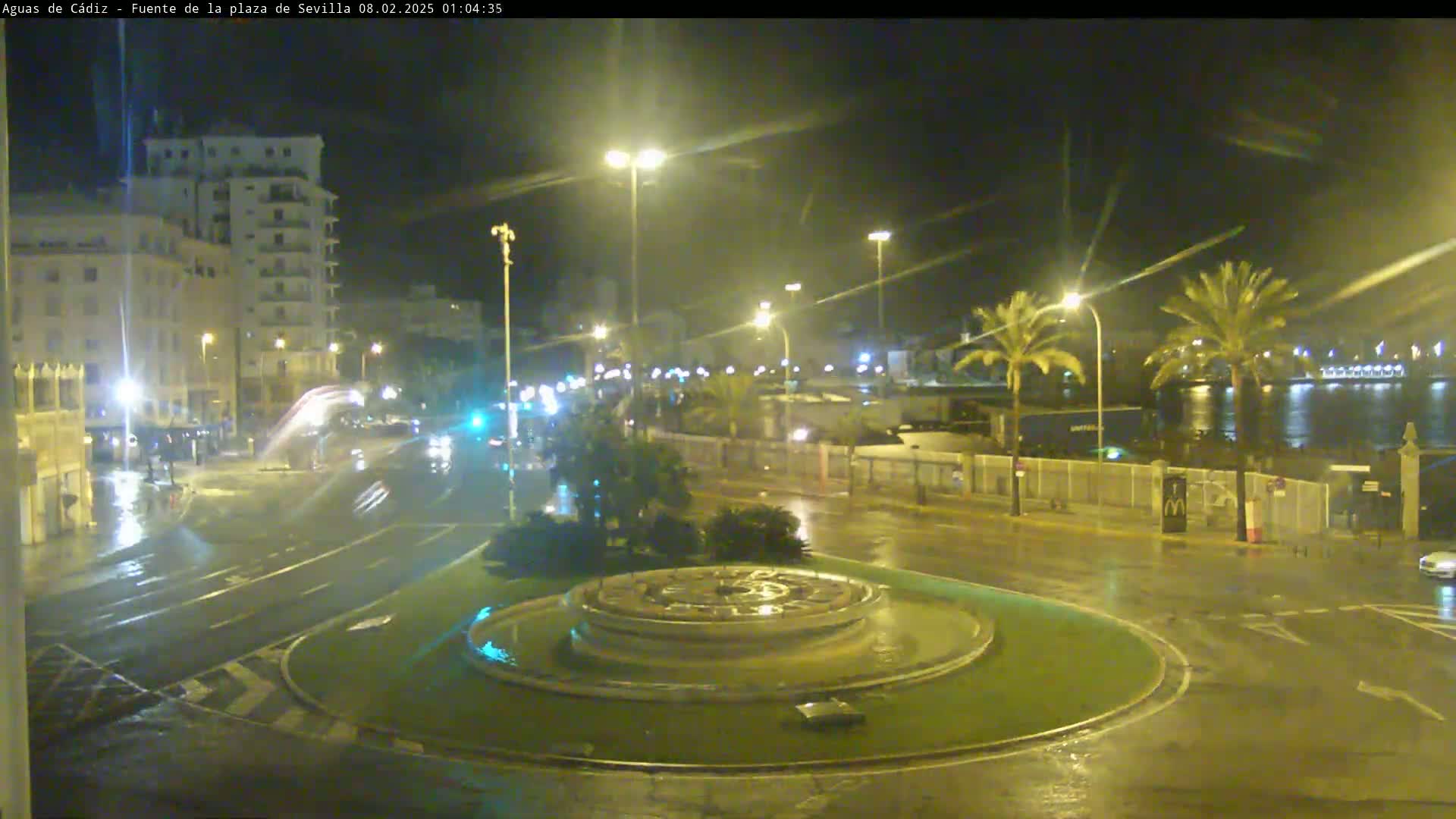 A nighttime, rain-slicked city scene shows a traffic circle with a fountain in the center, surrounded by buildings, palm trees, and streetlights.