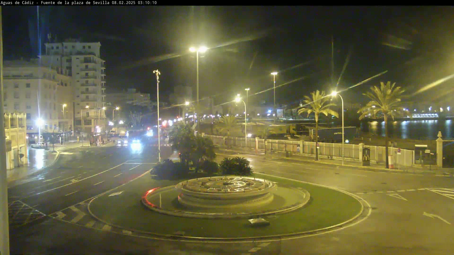 A nighttime view of a wet city roundabout with a fountain at its center, surrounded by buildings, palm trees, and streetlights, next to a calm body of water.