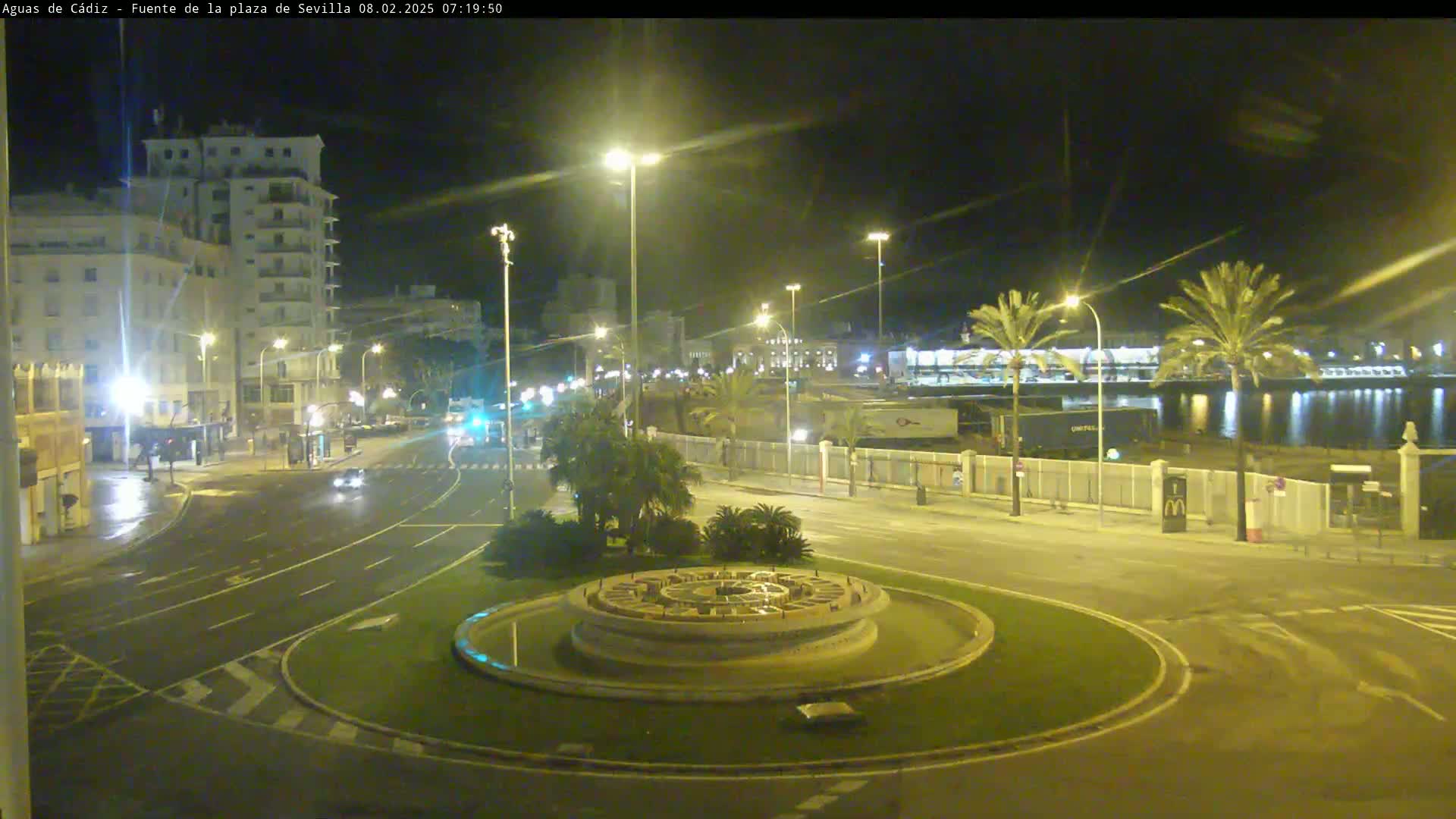 A nighttime view of a roundabout with a fountain in the center, surrounded by streets, buildings, and palm trees near a body of water under clear skies.