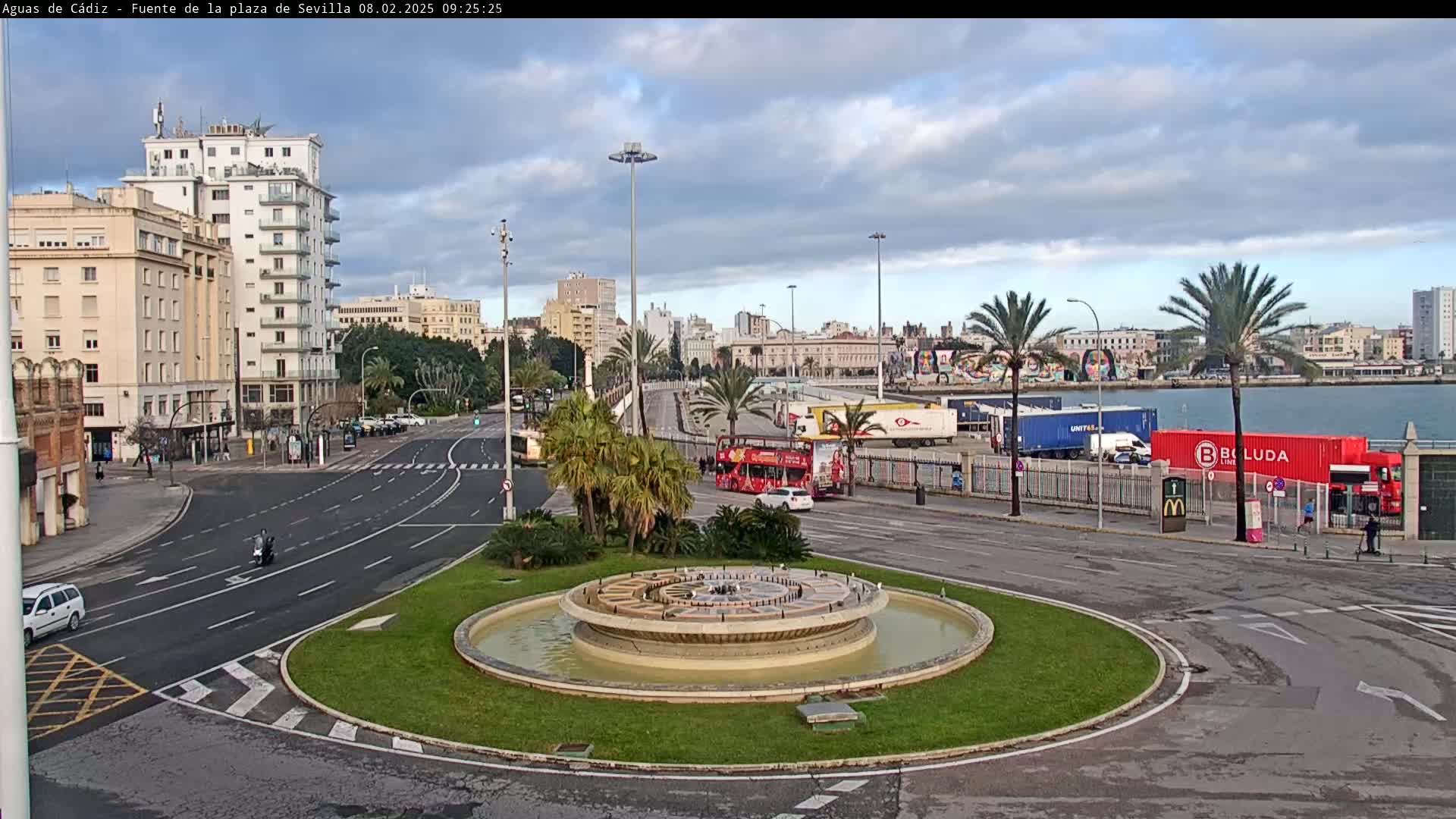 A circular fountain sits in a traffic circle next to a waterfront area with palm trees under a partly cloudy sky.