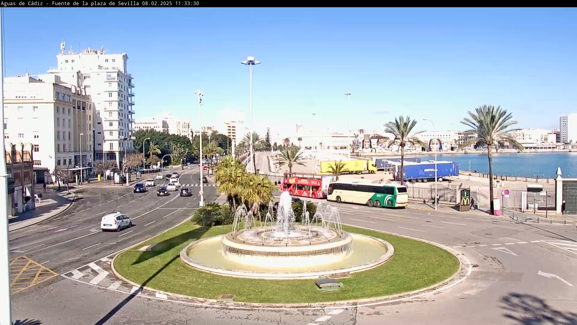 A circular fountain is at the center of a traffic circle, surrounded by palm trees, buildings, and buses, under a clear blue sky.
