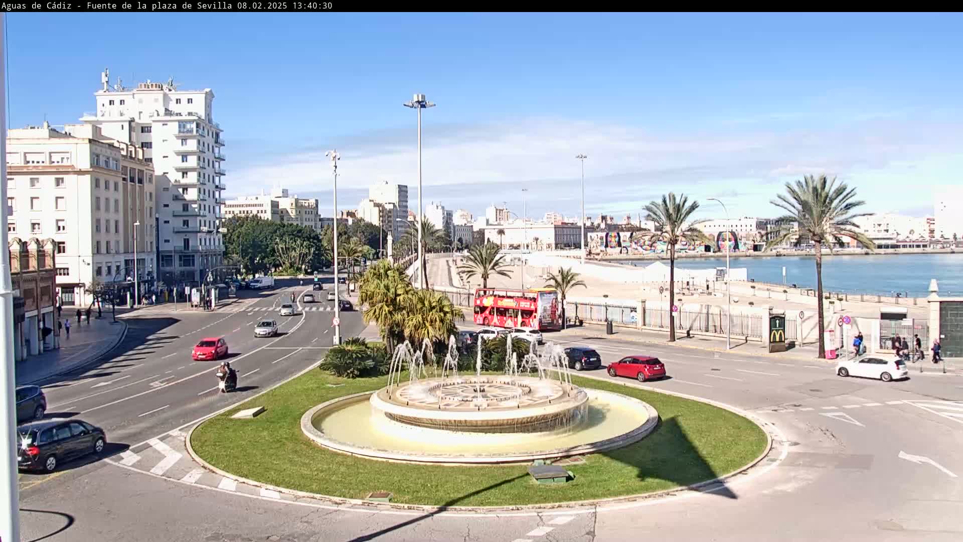 A circular fountain is in the center of a traffic circle, with buildings, palm trees, a double-decker bus, and cars visible, all under a clear sunny sky.
