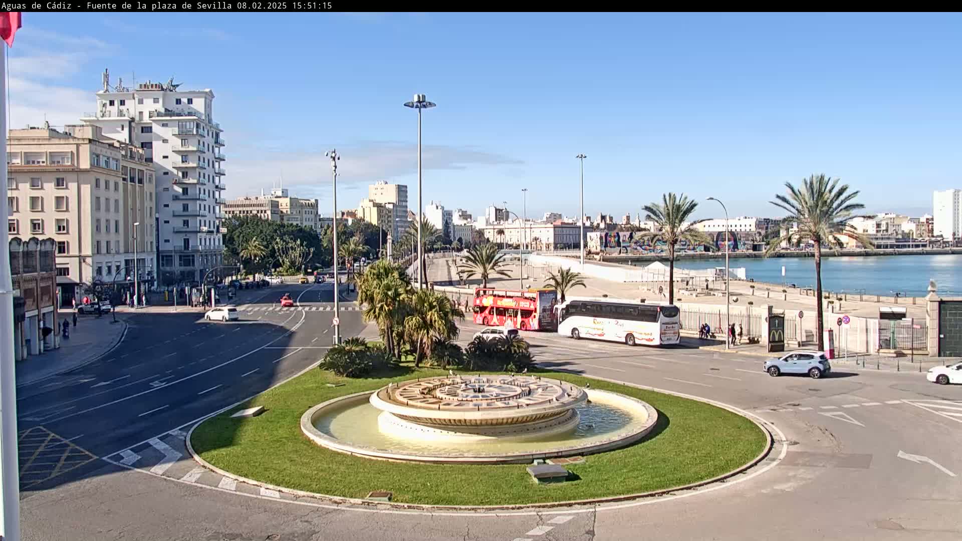 A circular fountain sits in the center of a traffic circle, surrounded by palm trees, buildings, and buses, under a clear blue sky.