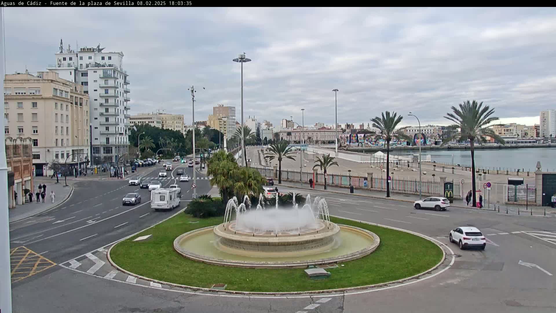 A circular fountain sprays water in the center of a traffic circle, surrounded by buildings, palm trees, cars, and a body of water under a cloudy sky.