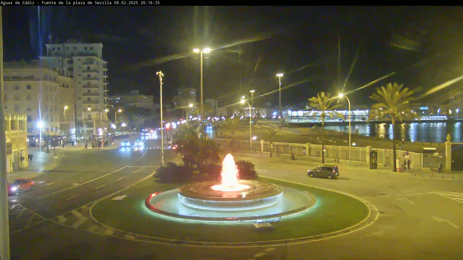 A nighttime view of a city square with a lit fountain at its center, surrounded by streets with cars and buildings, and a waterfront in the background.