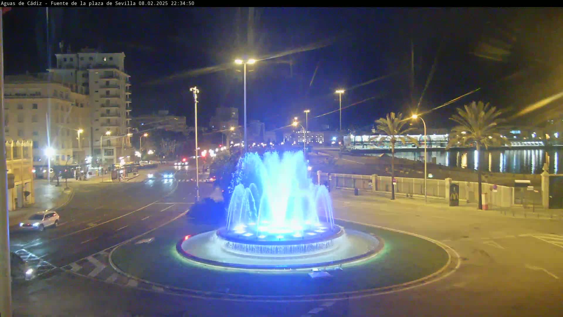 A nighttime view of a city square featuring a large illuminated fountain in the center, surrounded by roads and buildings, with palm trees and water visible in the background.