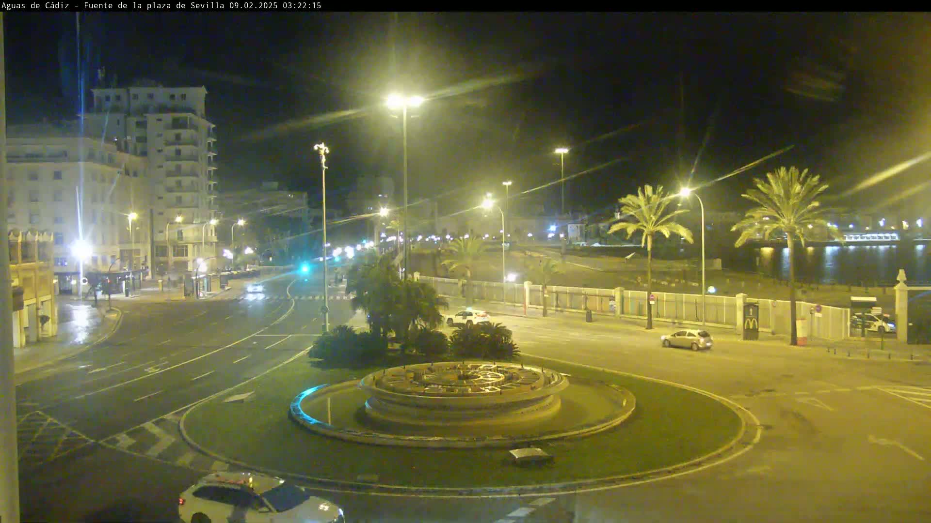 A nighttime view of a city roundabout with a fountain at its center, wet roads, and palm trees lining a waterfront in the background.