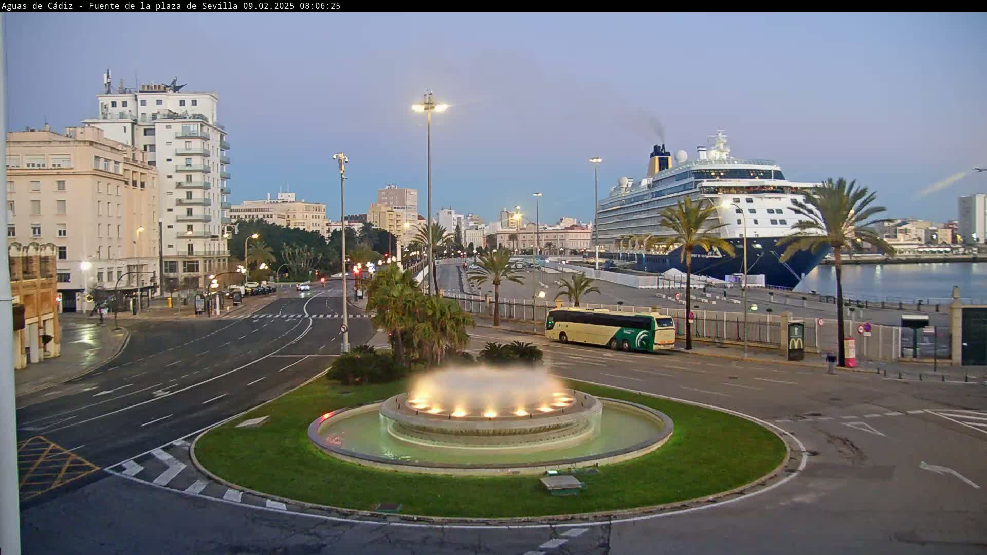 A large cruise ship is docked at a port in a city with a circular fountain in the foreground, under a twilight sky.