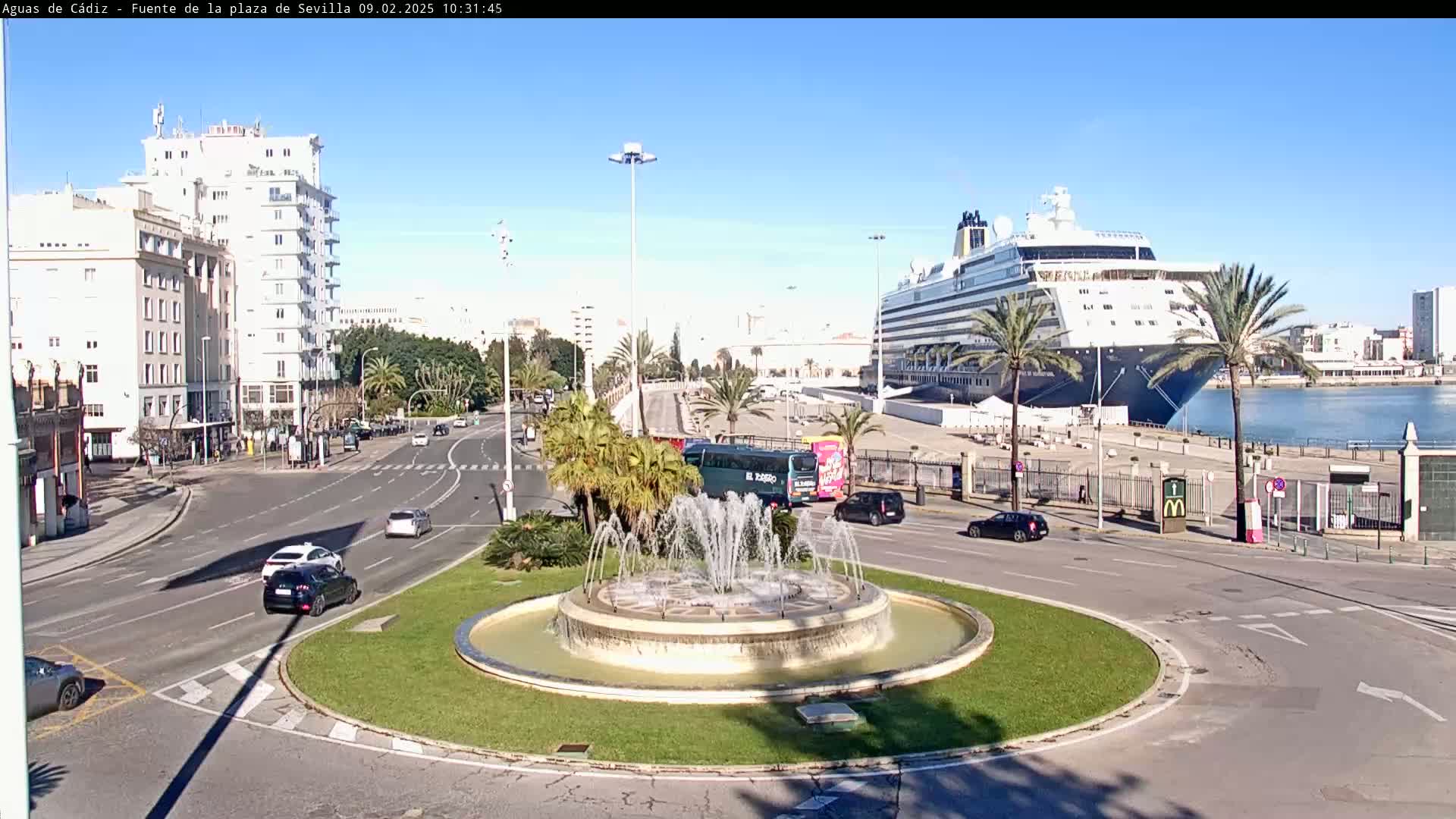 A large cruise ship is docked at a port, next to a roundabout with a fountain, surrounded by buildings and palm trees under a clear sunny sky.