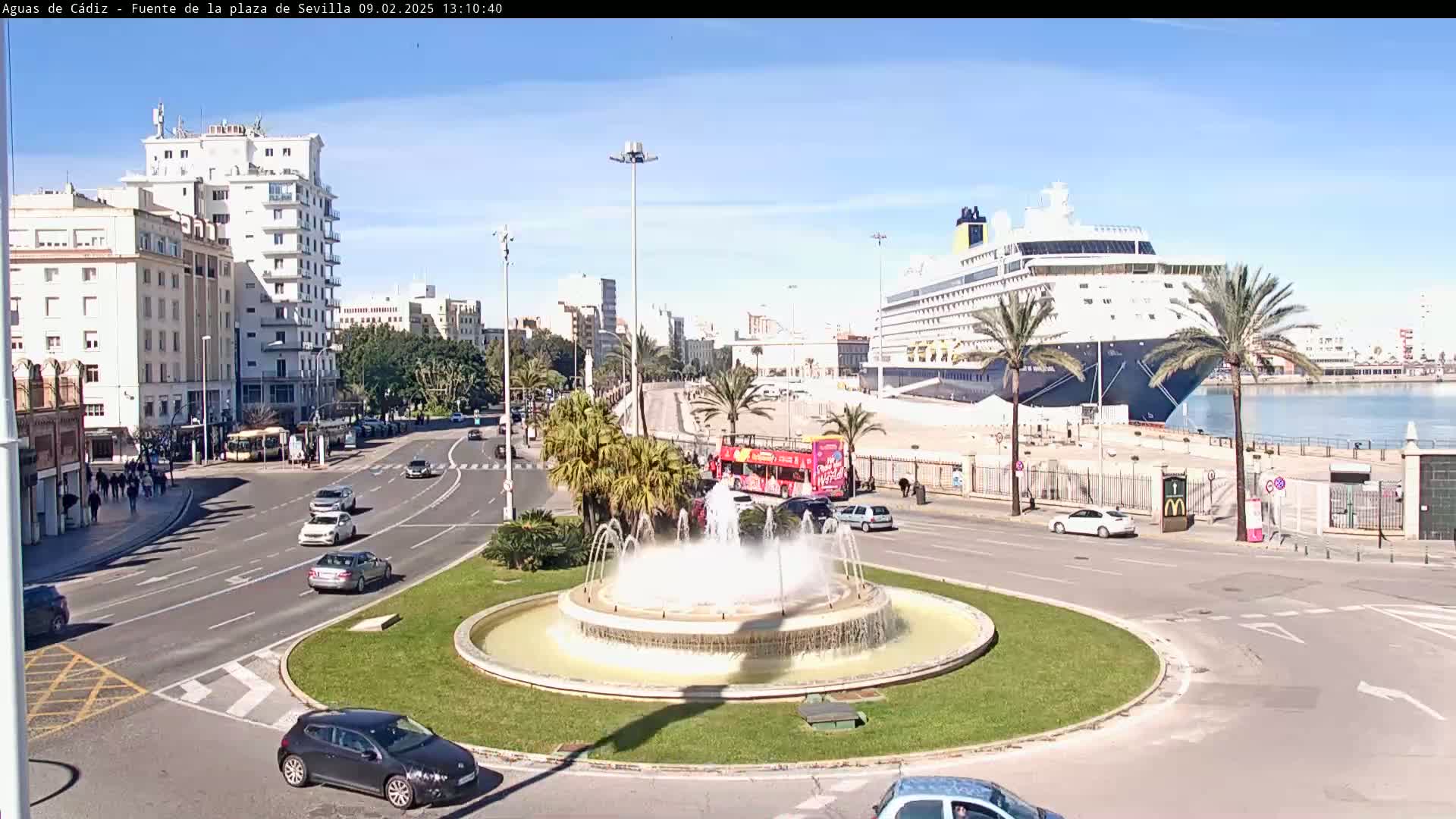 A large cruise ship is docked at a port, near a roundabout with a fountain and city buildings under a clear, sunny sky.
