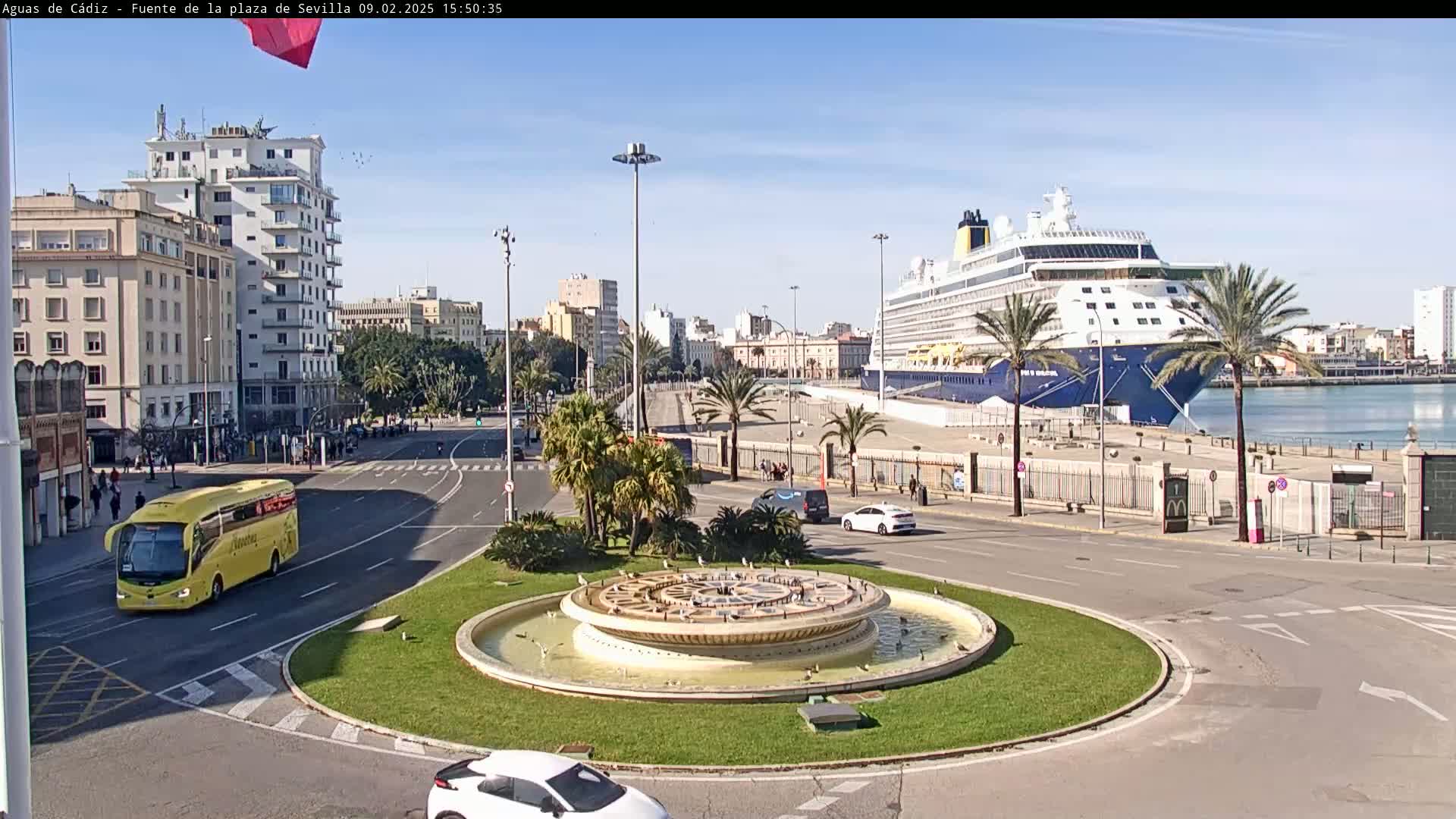 A large cruise ship is docked at a port city on a sunny day, with a circular fountain and a yellow bus visible in the foreground.