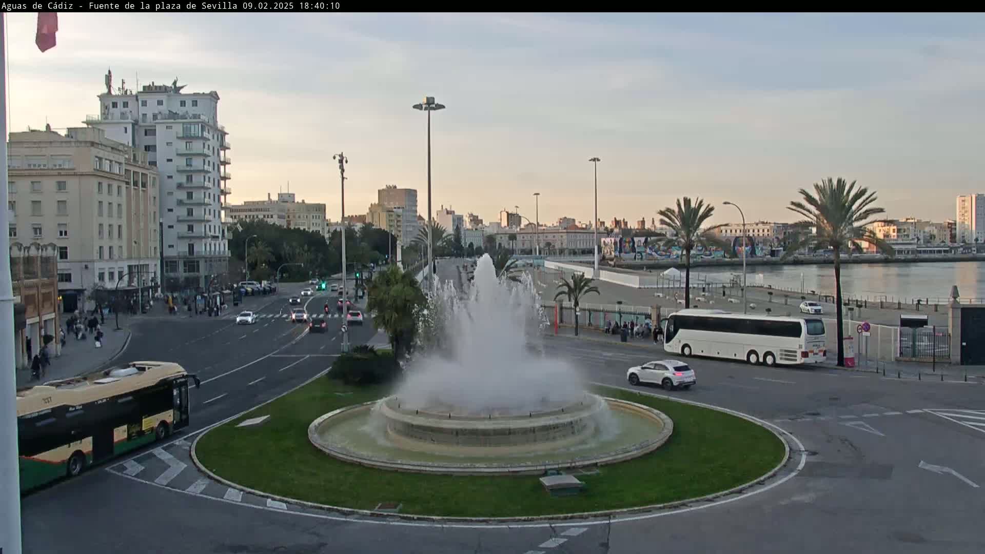 A large fountain in a city roundabout sprays water under a partly cloudy evening sky, with buses, cars, and buildings visible in the background.