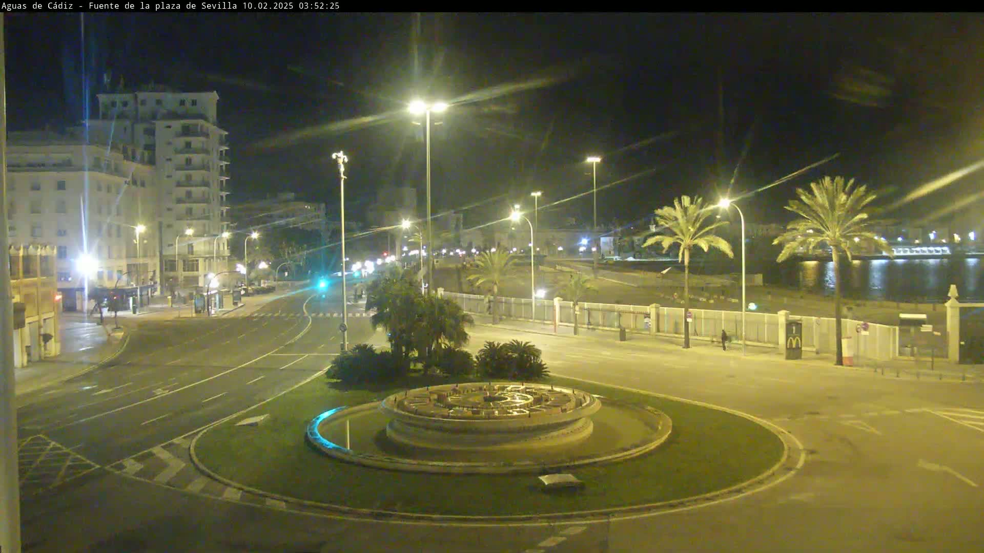 A nighttime view of a city roundabout with a fountain at its center, illuminated by streetlights, with palm trees and buildings in the background near a body of water.