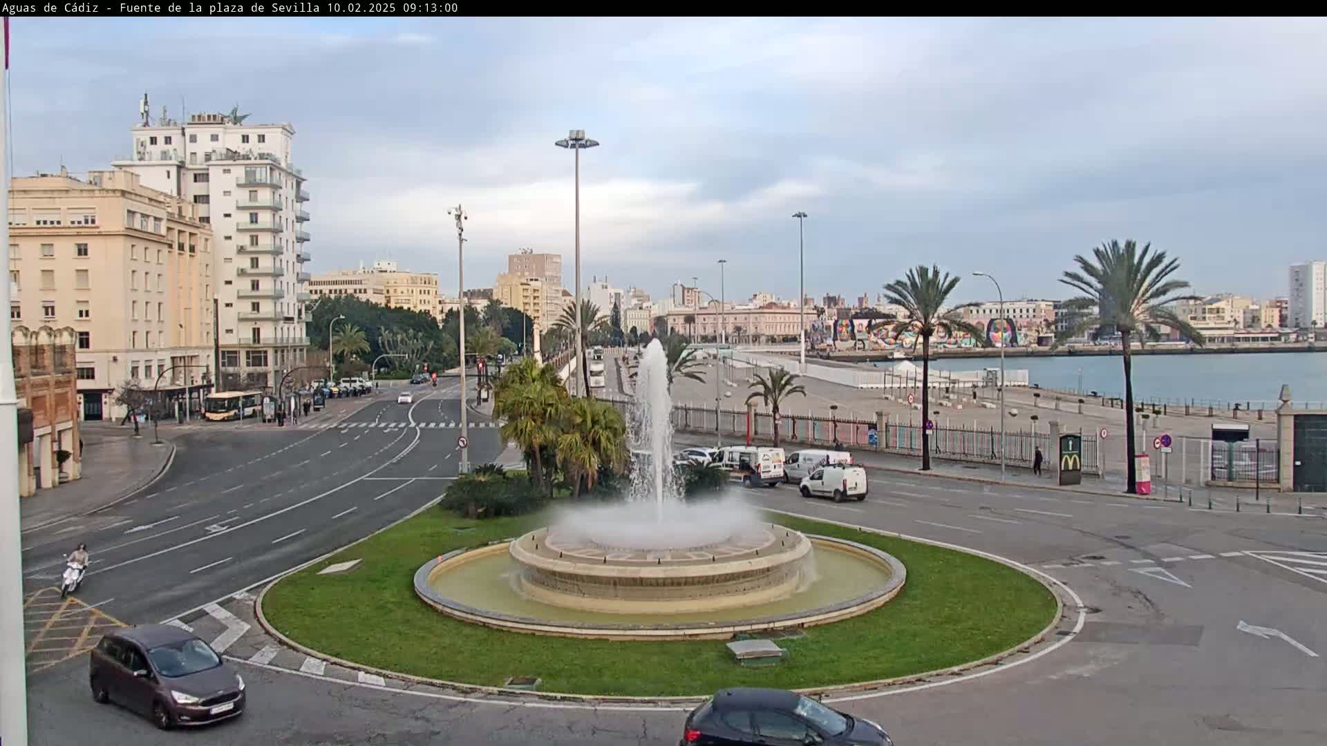 A large fountain in a city roundabout is surrounded by palm trees, buildings, and roads, under a mostly cloudy sky.