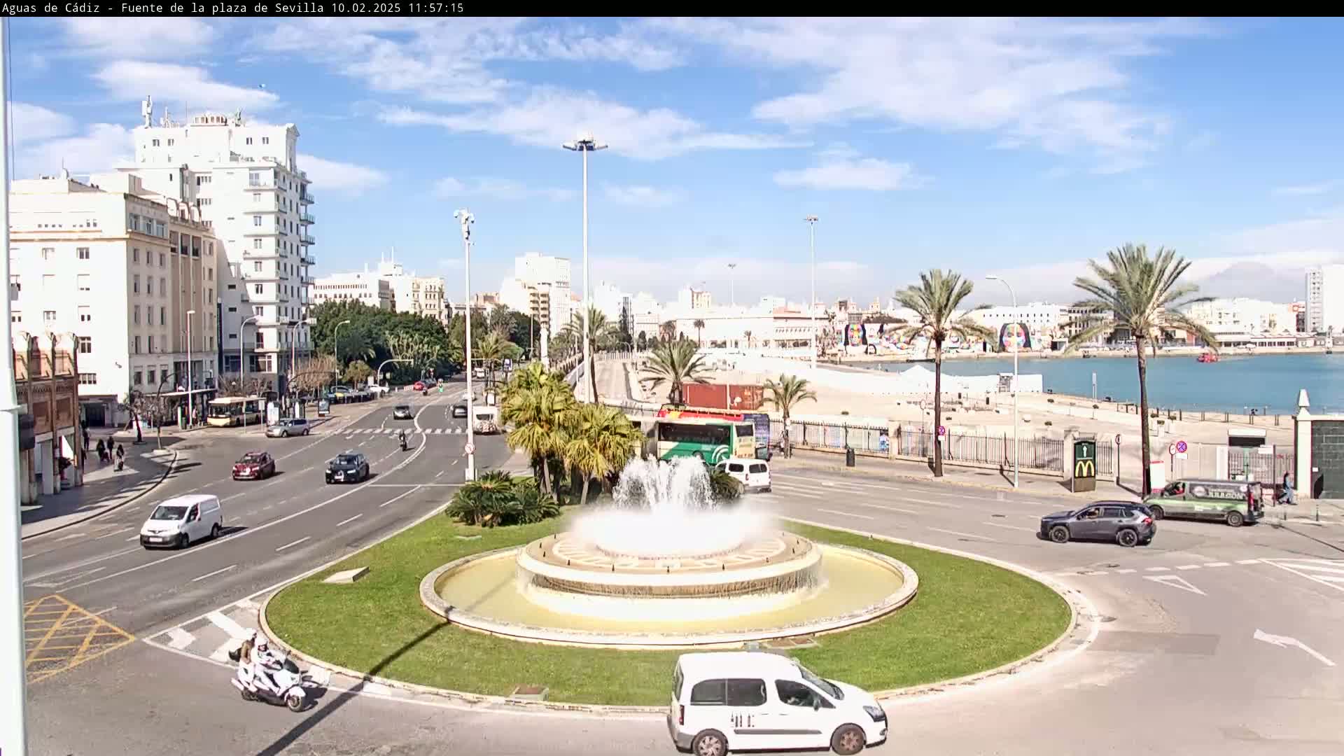 A city roundabout with a fountain in the center, surrounded by palm trees, buildings, and traffic, under a partly cloudy sky.