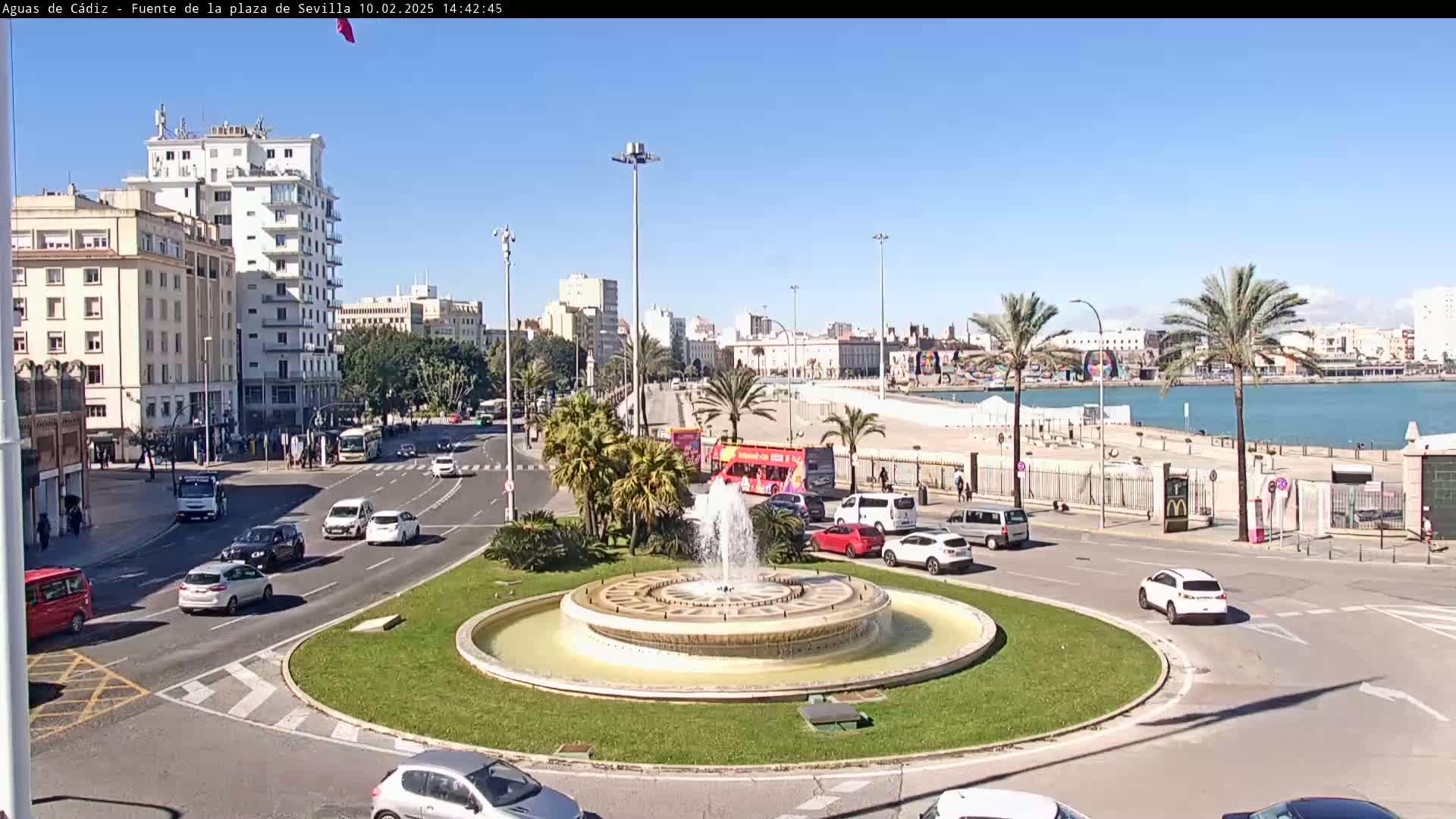 A sunny day reveals a city scene centered around a circular fountain in a traffic circle, with buildings, palm trees, cars, and a body of water in the distance.