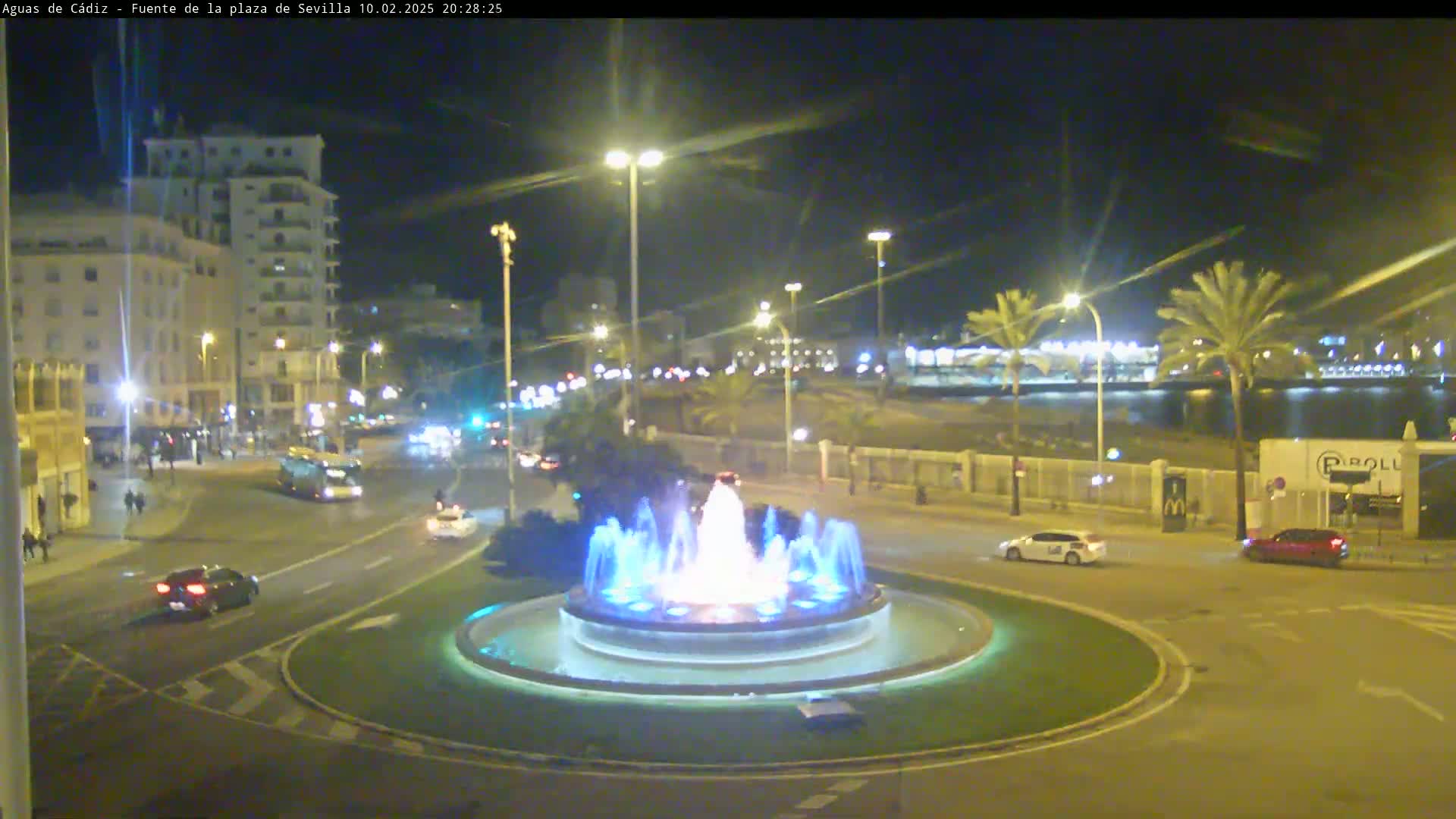 A nighttime view of a city roundabout featuring a lit fountain, with cars driving around it and buildings and palm trees visible in the background under a clear sky.