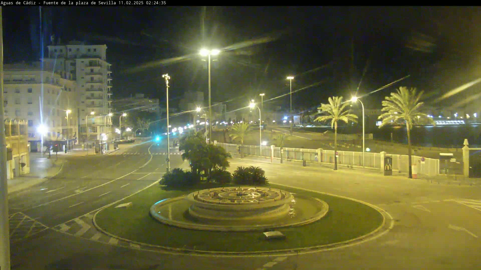 A nighttime view of a circular traffic island with a fountain in the center, surrounded by streets, buildings, and palm trees under clear skies.