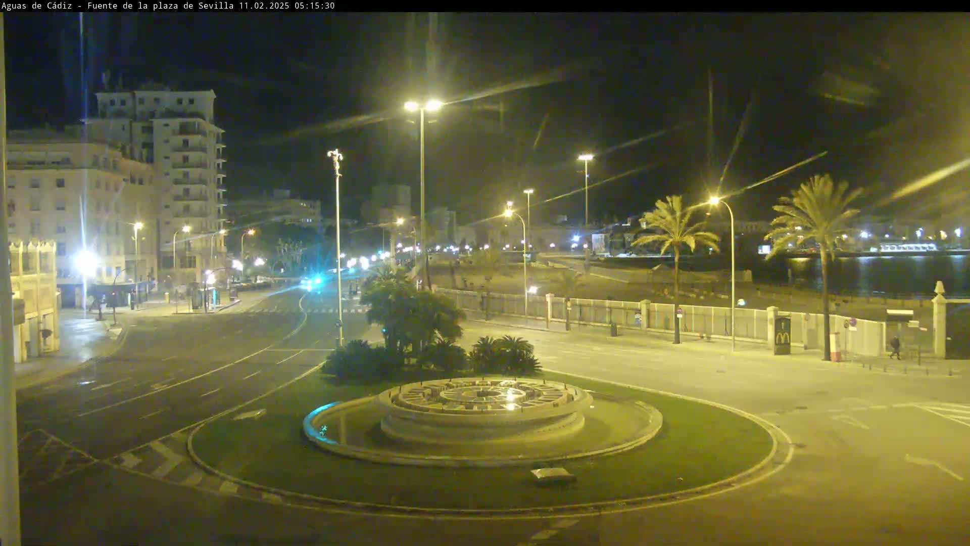 A nighttime view of a city roundabout with a circular fountain at its center, surrounded by palm trees, buildings, and streetlights, near a body of water under a clear sky.