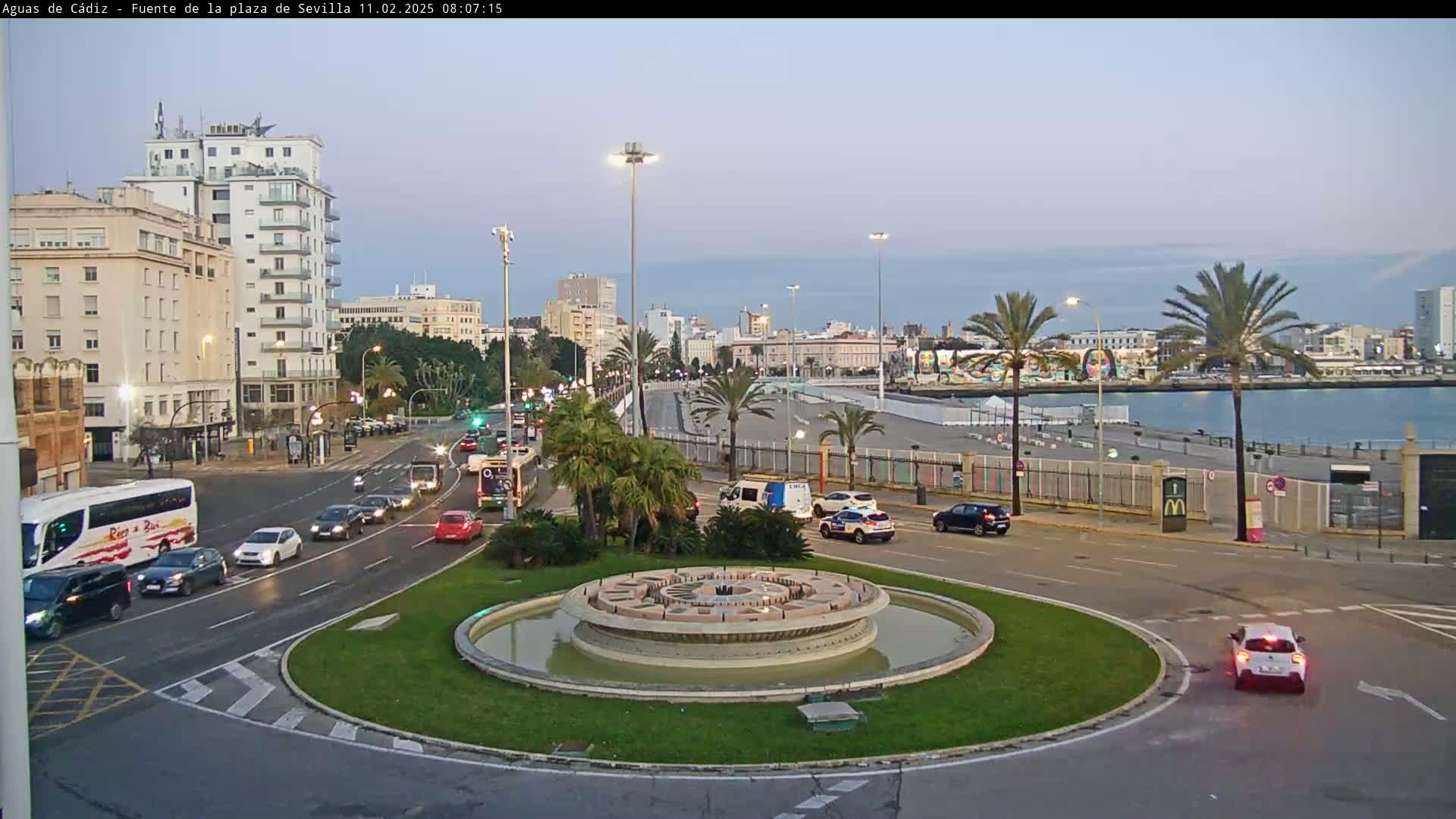 A city roundabout with a central fountain is surrounded by palm trees, buildings, and cars under a twilight sky.