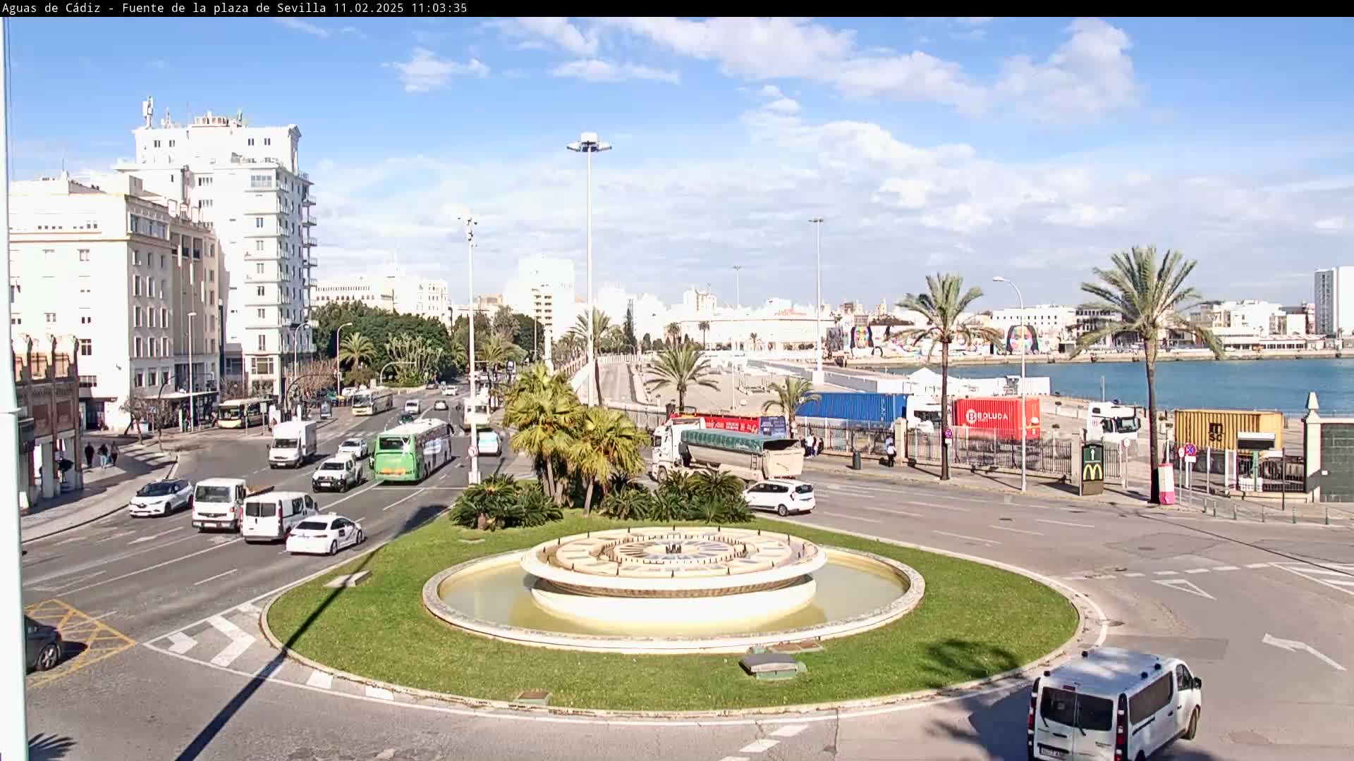 A roundabout with a central fountain is surrounded by city streets with cars and buses under a mostly sunny sky.