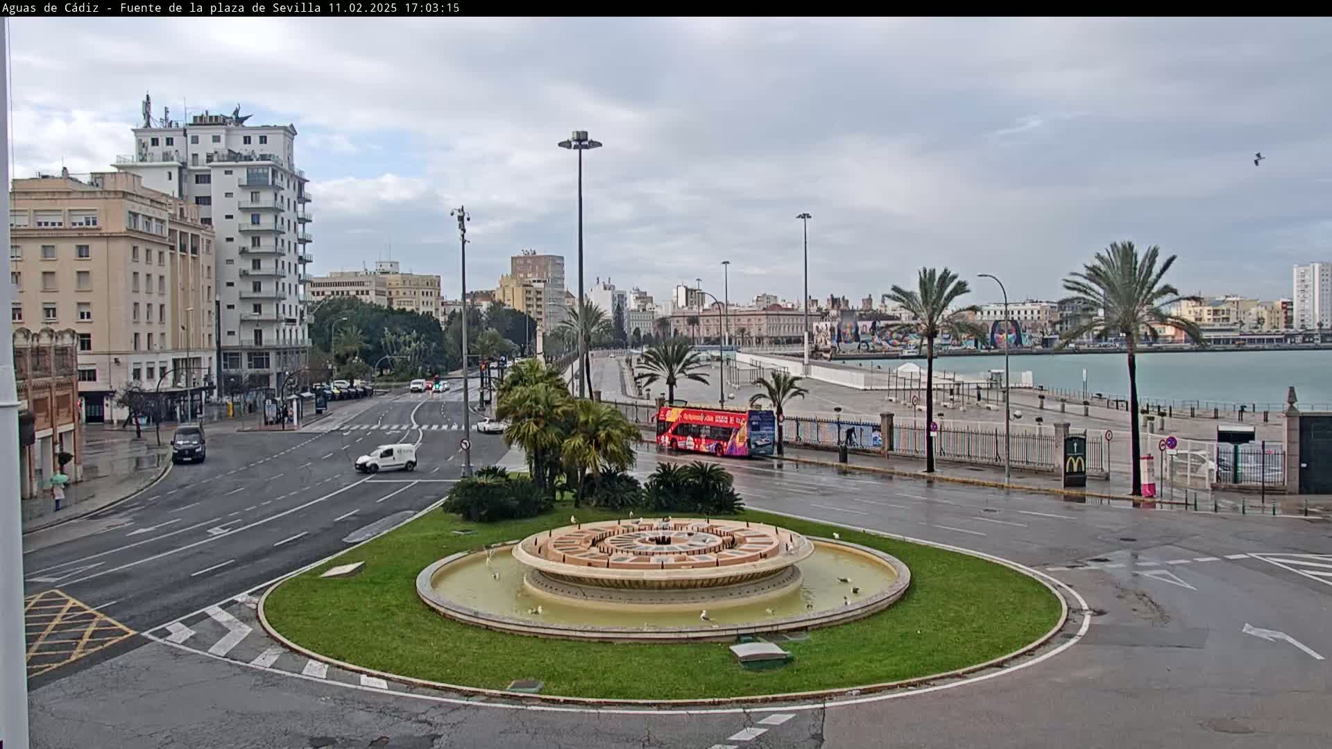 A wet city square with a circular fountain in the center, surrounded by roads, buildings, palm trees, and a waterfront in the background under an overcast sky.