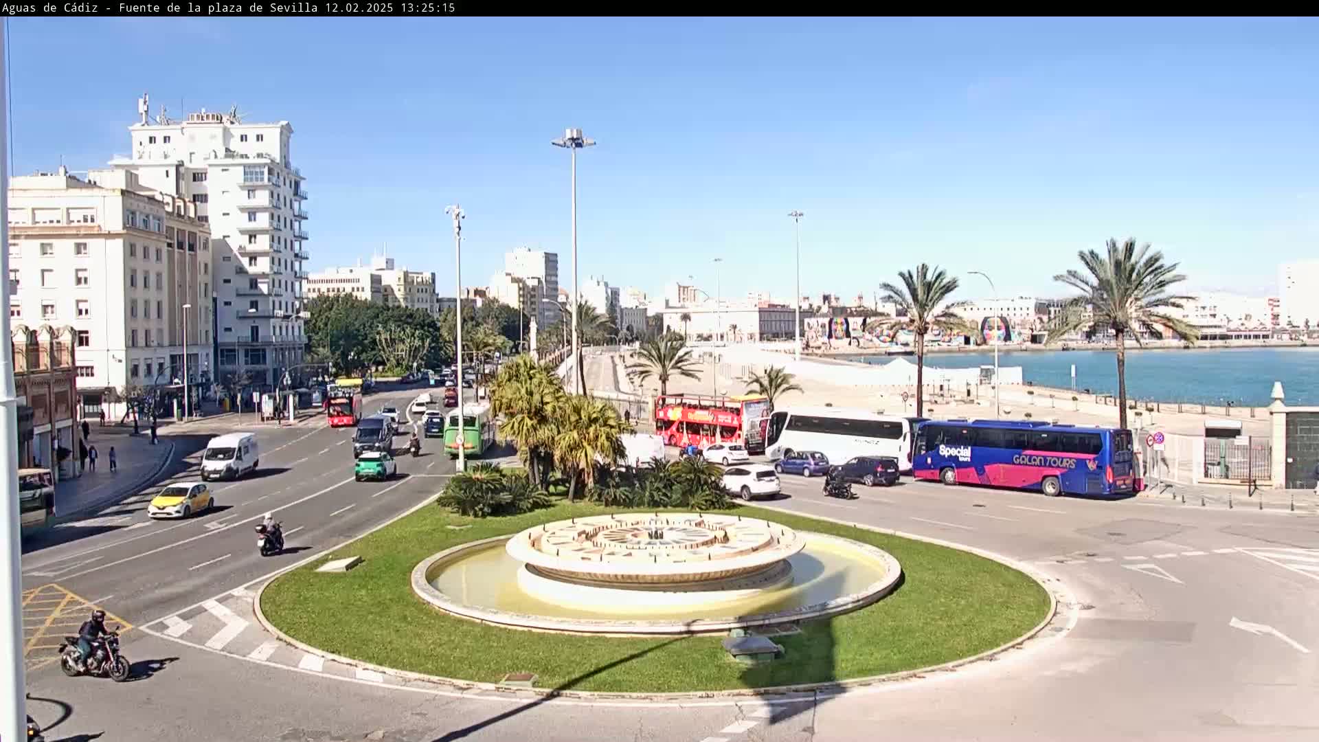 A circular fountain sits in the center of a traffic circle, surrounded by palm trees, buildings, and various vehicles on a sunny day.