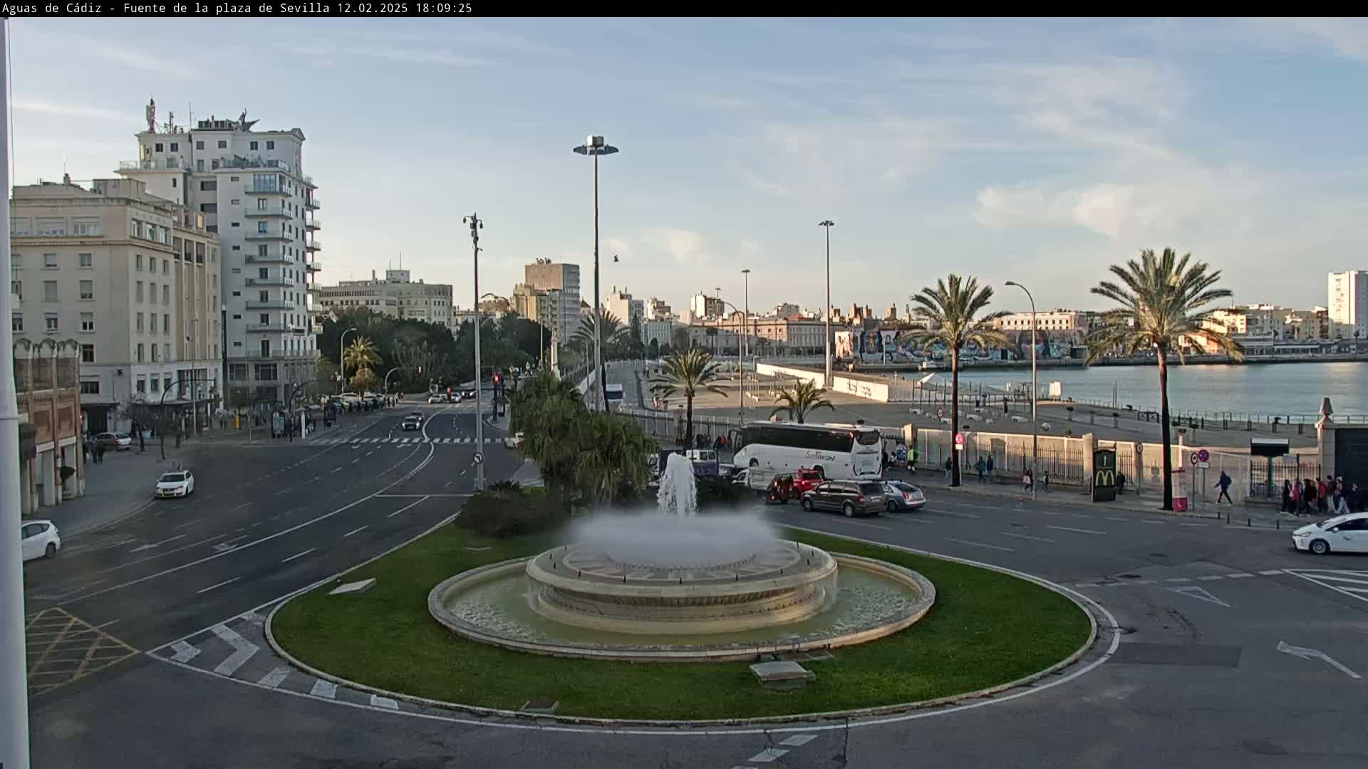 A city square with a circular fountain at its center, surrounded by roads, buildings, and palm trees, under a partly cloudy sky.