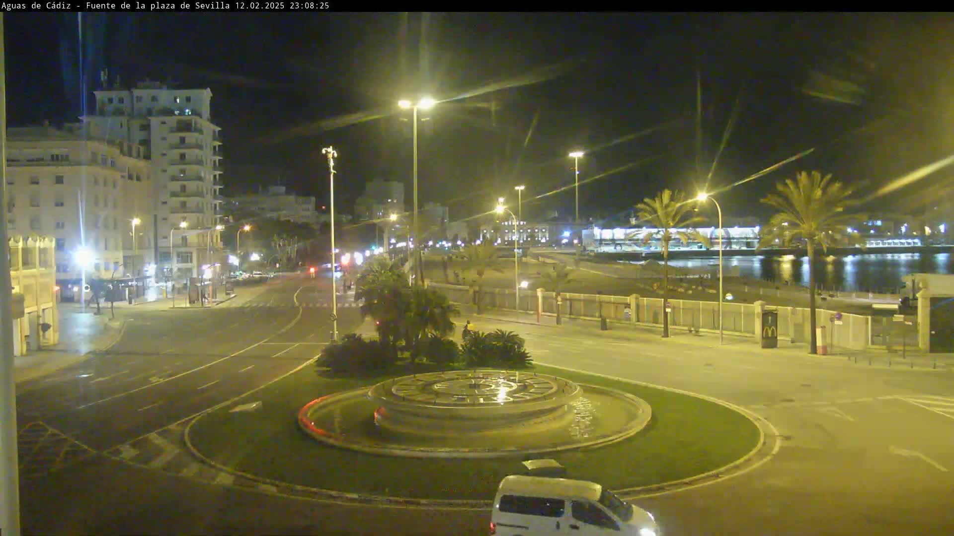 A nighttime view of a city roundabout with a fountain at its center, showing a waterfront area with palm trees and buildings under clear skies.