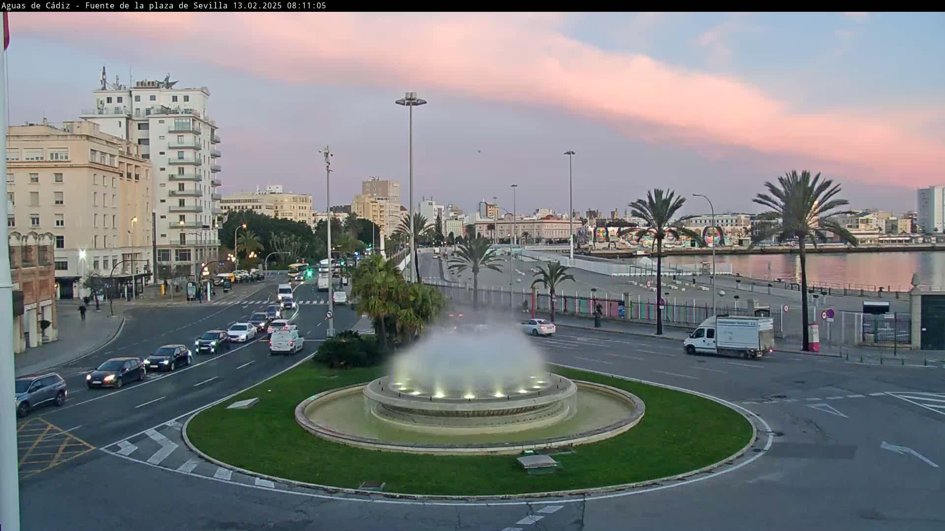 A city fountain with a misty spray is situated in a traffic circle, with city buildings and a waterfront visible in the background under a pinkish-orange sunset sky.