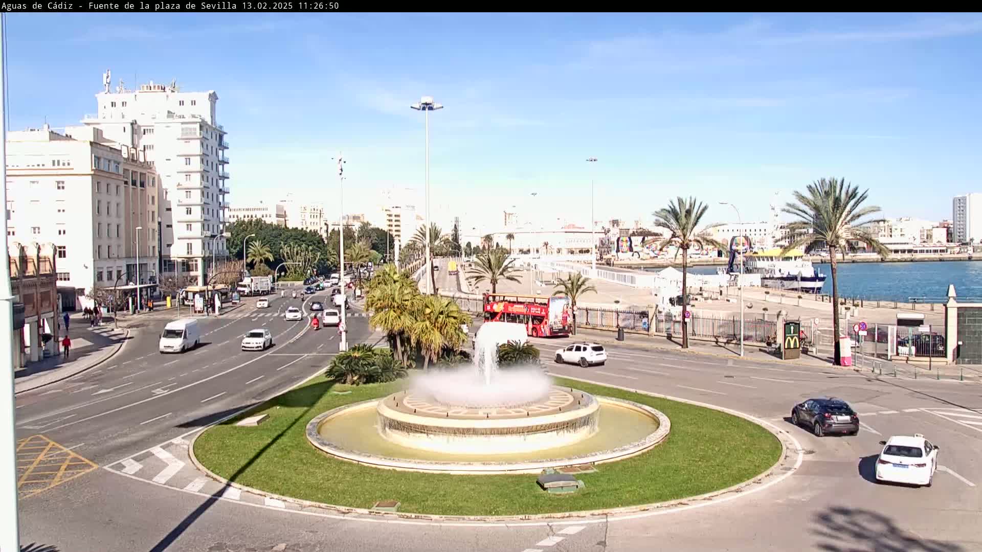 A city roundabout with a central fountain, surrounded by palm trees, buildings, and light traffic, under a clear sunny sky.