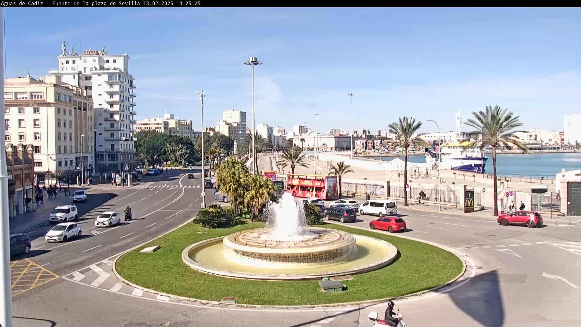 A city roundabout with a fountain in the center, surrounded by buildings, palm trees, cars, and a double-decker bus, on a sunny day near a harbor with a ship.