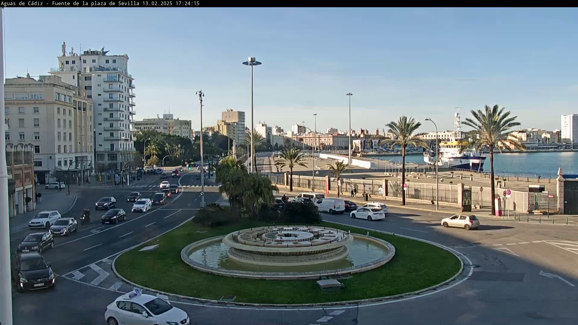 A city roundabout with a fountain in the center, surrounded by buildings and cars, overlooks a harbor with a docked ship under a clear, sunny sky.