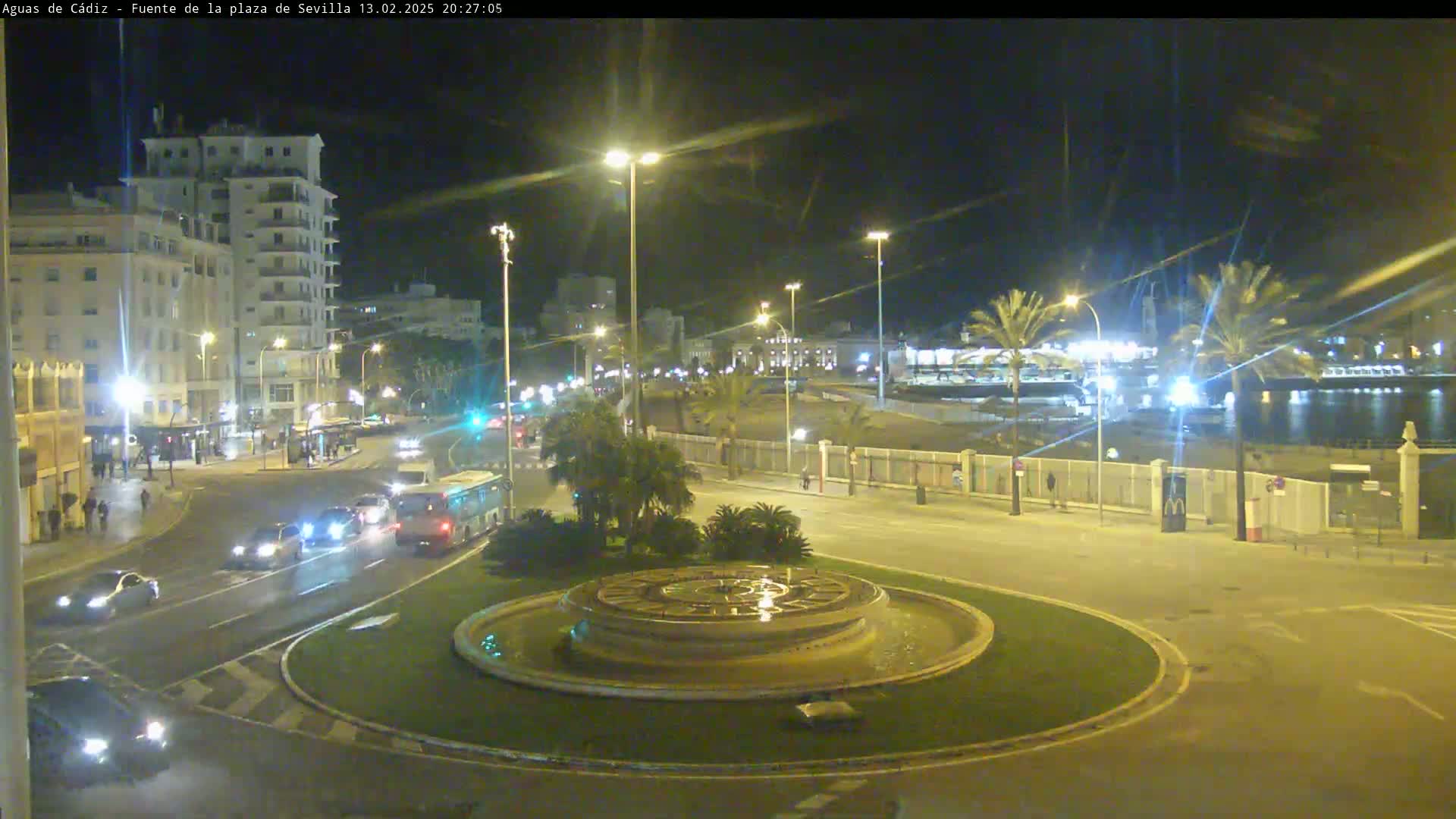 A nighttime view of a city intersection with a circular fountain in the center, featuring traffic, buildings, palm trees, and a waterfront in the background under clear skies.