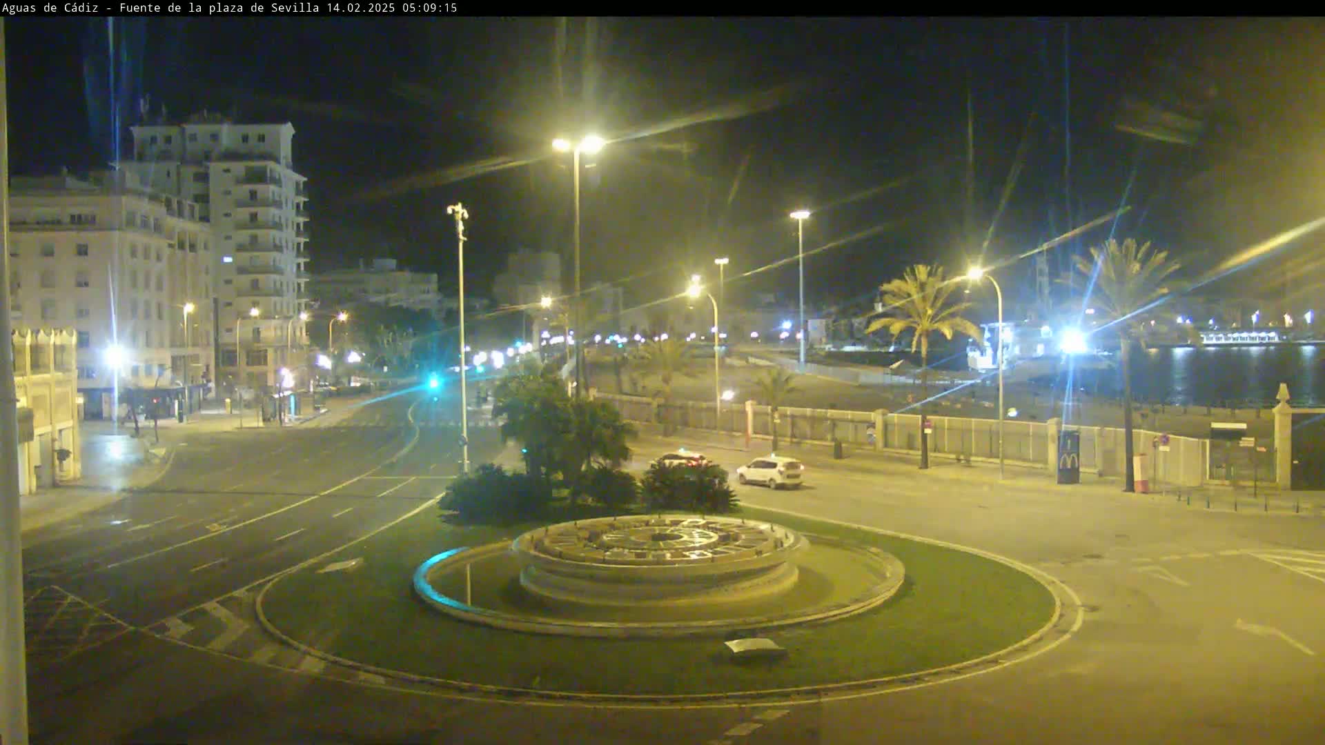 A nighttime view of a city roundabout with a fountain at its center,  streetlights illuminating buildings and palm trees along a waterfront road.